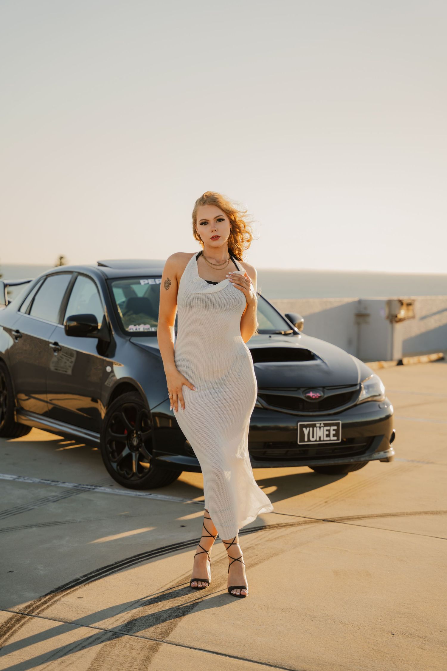 Blonde woman in white bodycon dress standing before black Subaru WRX at golden hour.