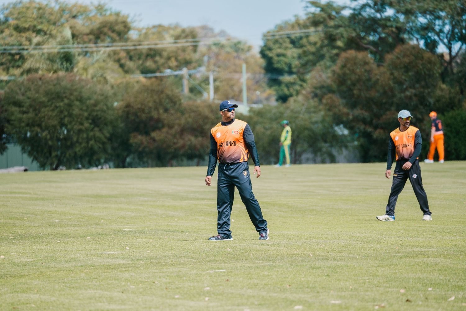 Two cricketers in orange uniforms stand in the outfield during a match.