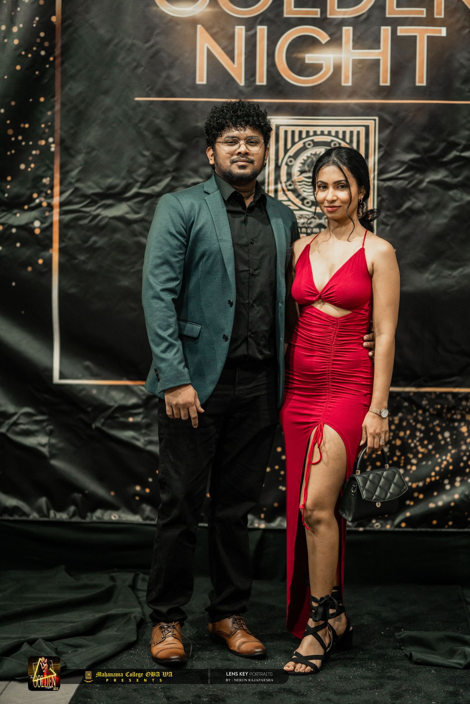A man in a teal suit and woman in a red dress posing at a Golden Night event backdrop.