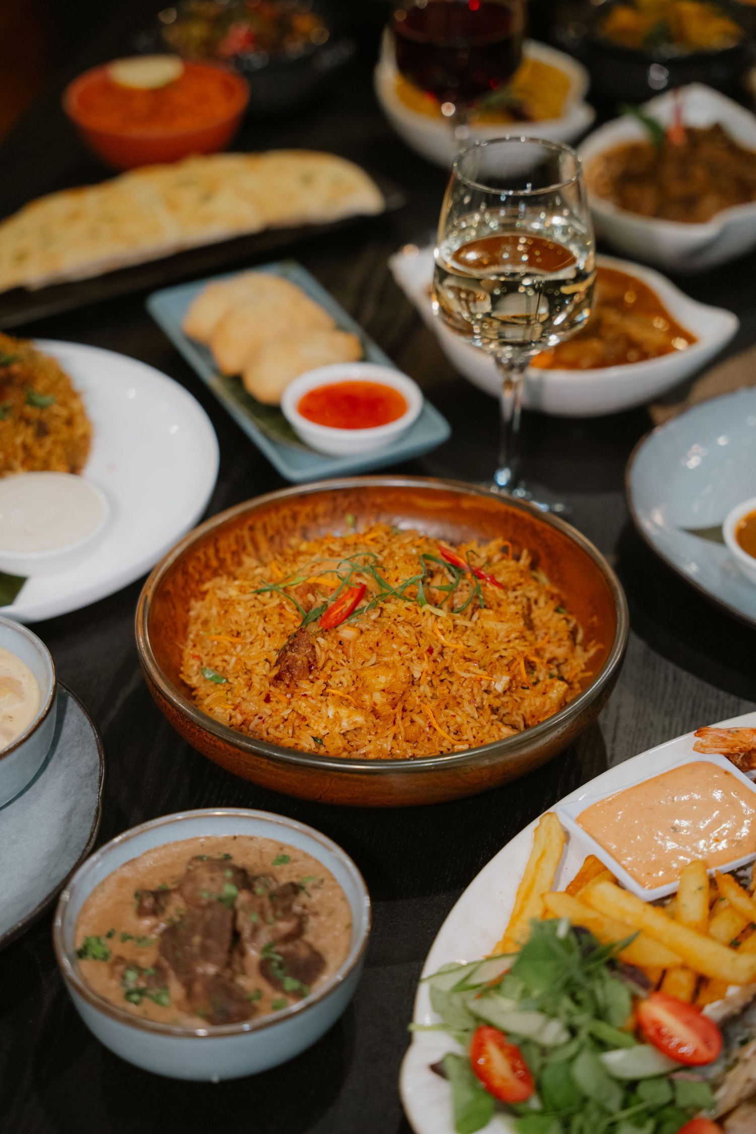 A large bowl of spiced rice surrounded by Indian dishes, fries, salad, and a glass of wine.