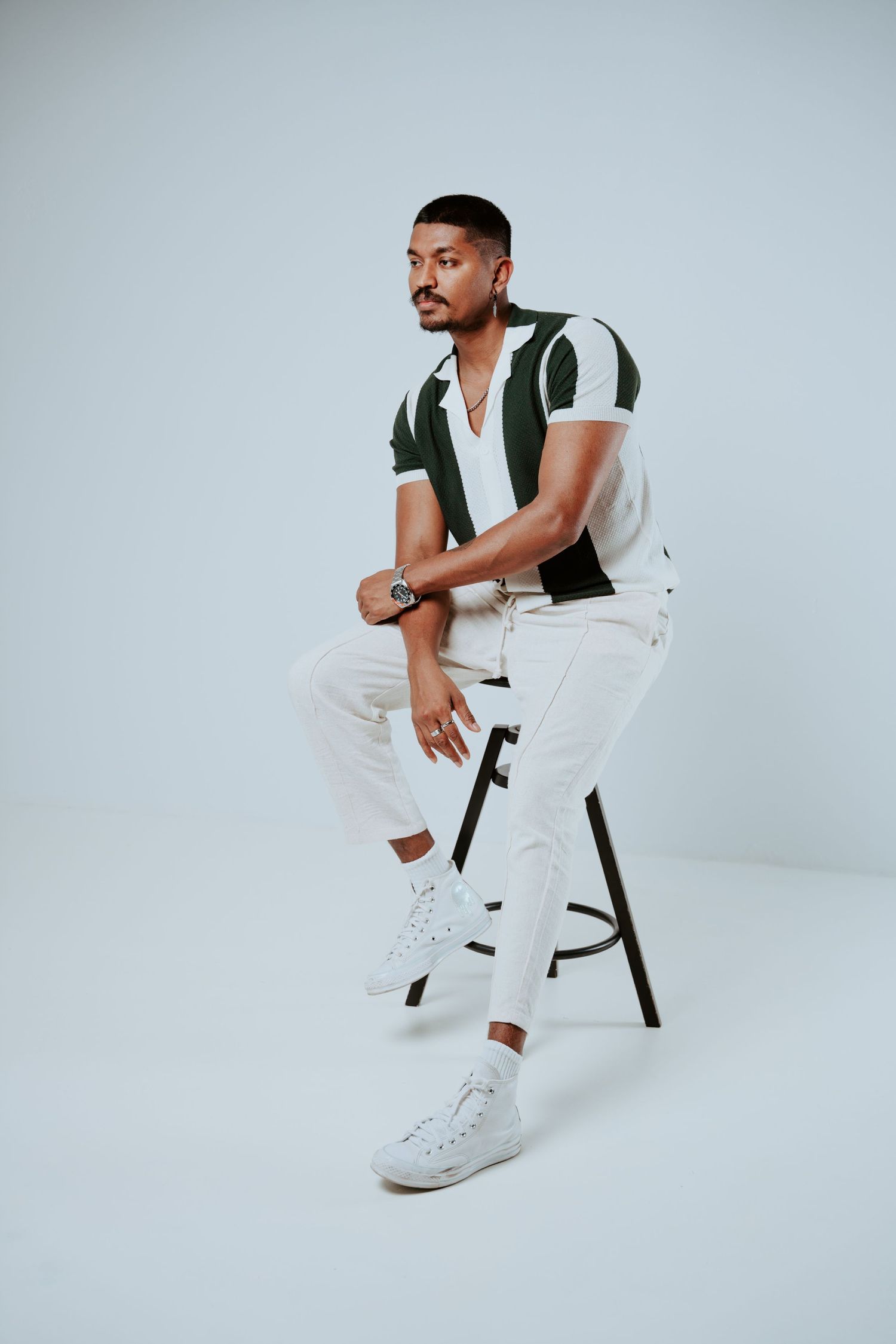 Stylish man sitting on stool wearing green striped polo shirt with white pants and sneakers against gray background.