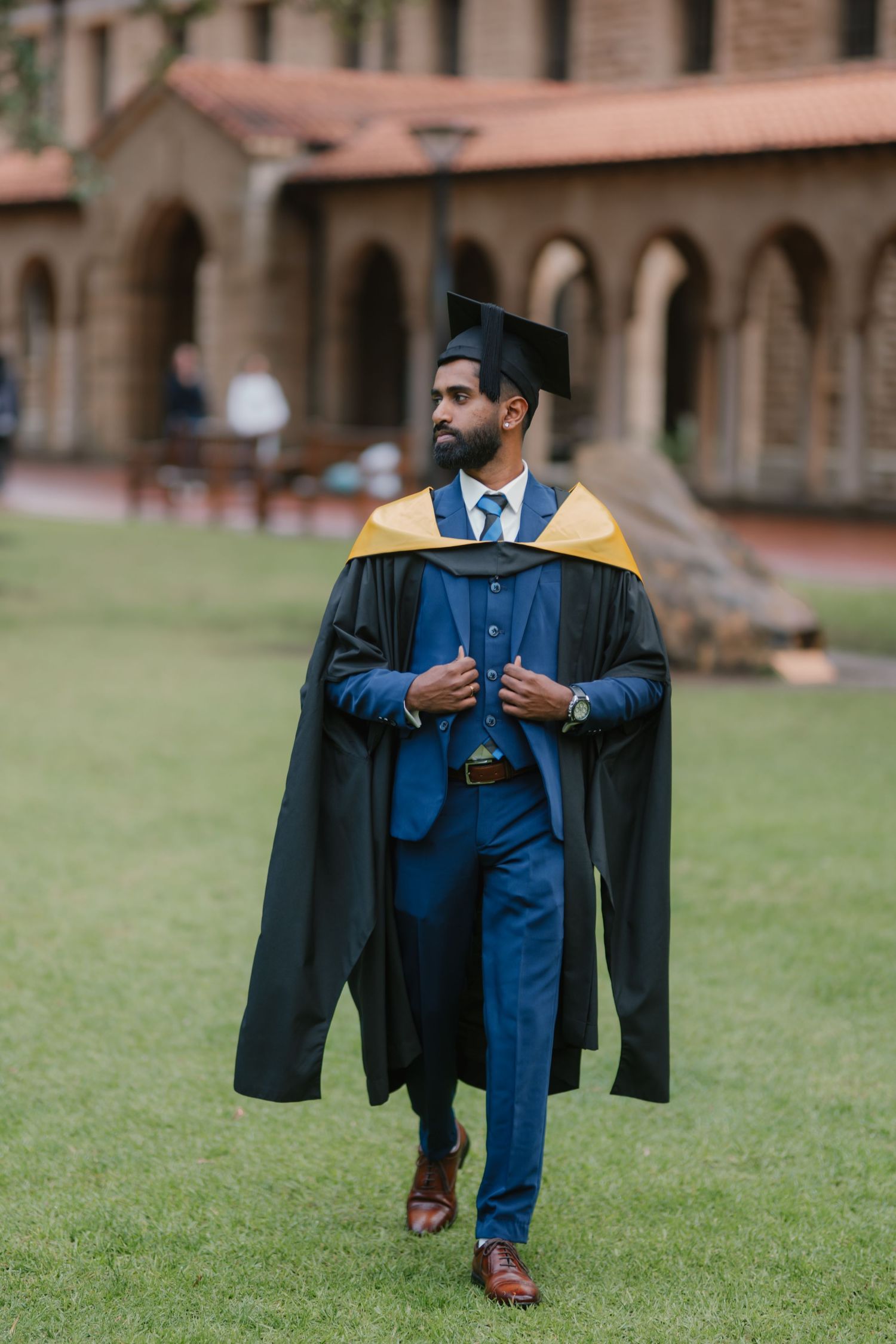 Male graduate in blue suit and black cap and gown walks across university campus lawn.