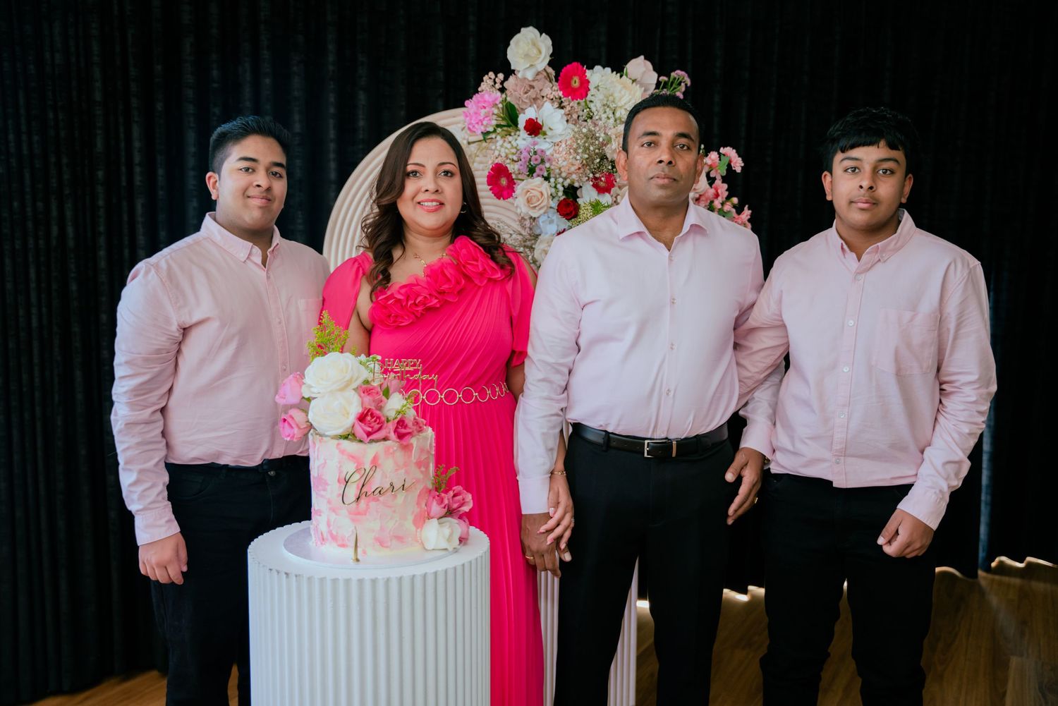 Family of four in pink outfits posing together beside a floral birthday cake at a celebration event.