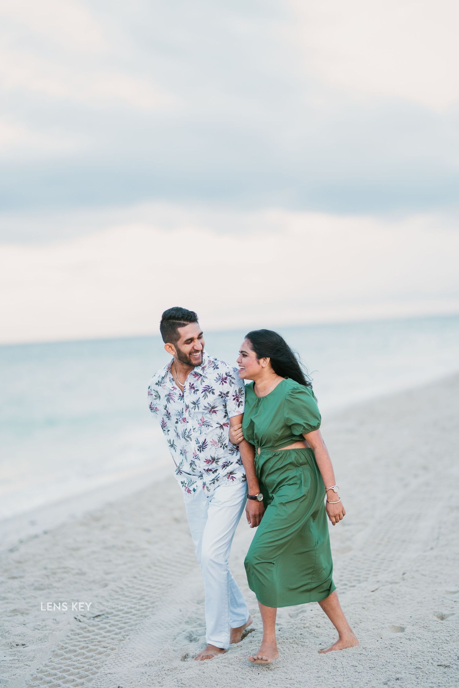 Couple walking hand in hand barefoot on a sandy beach, woman in green dress and man in floral shirt.