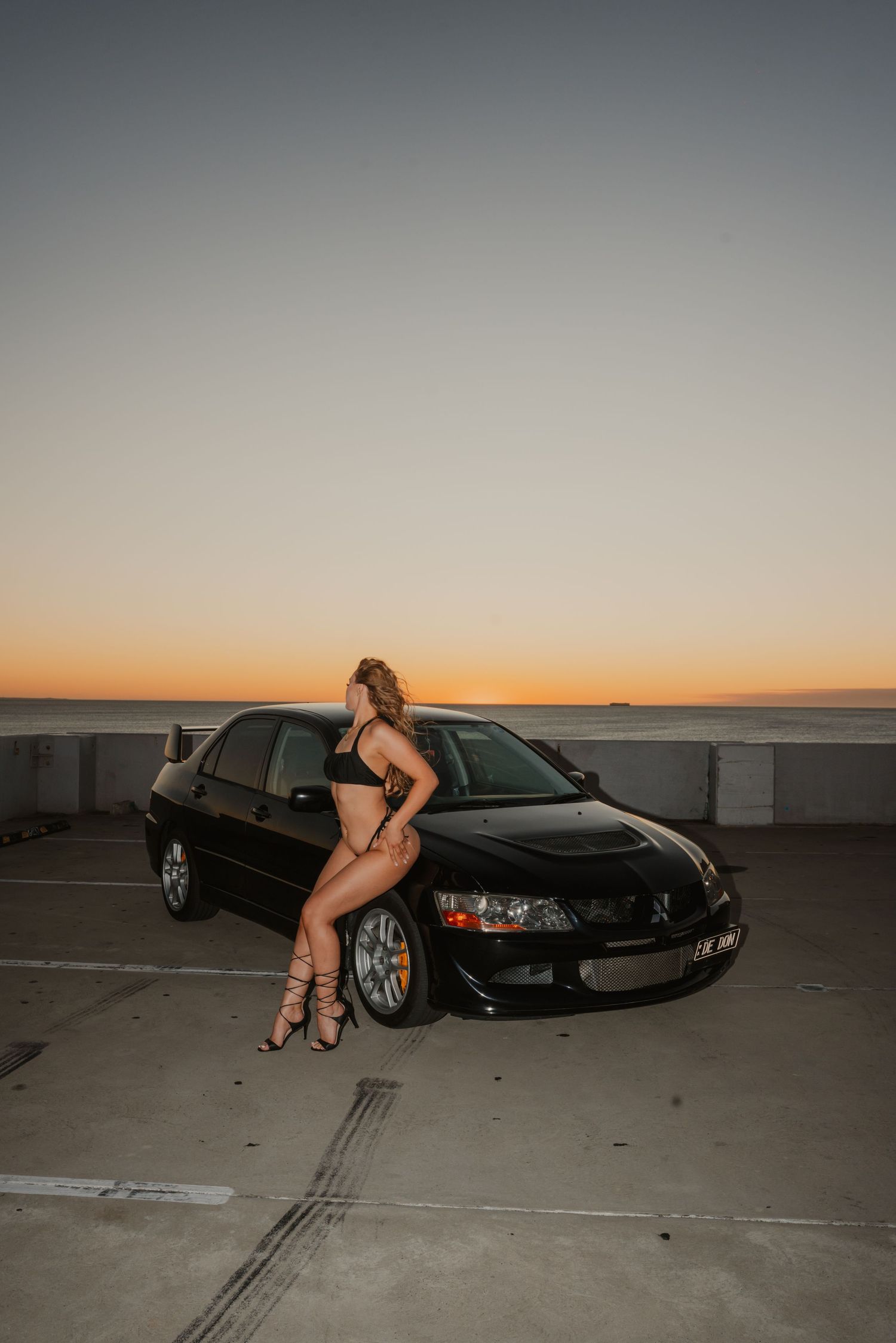 Woman in black swimwear leaning on a black Mitsubishi Evo sedan during a golden hour sunset photoshoot.