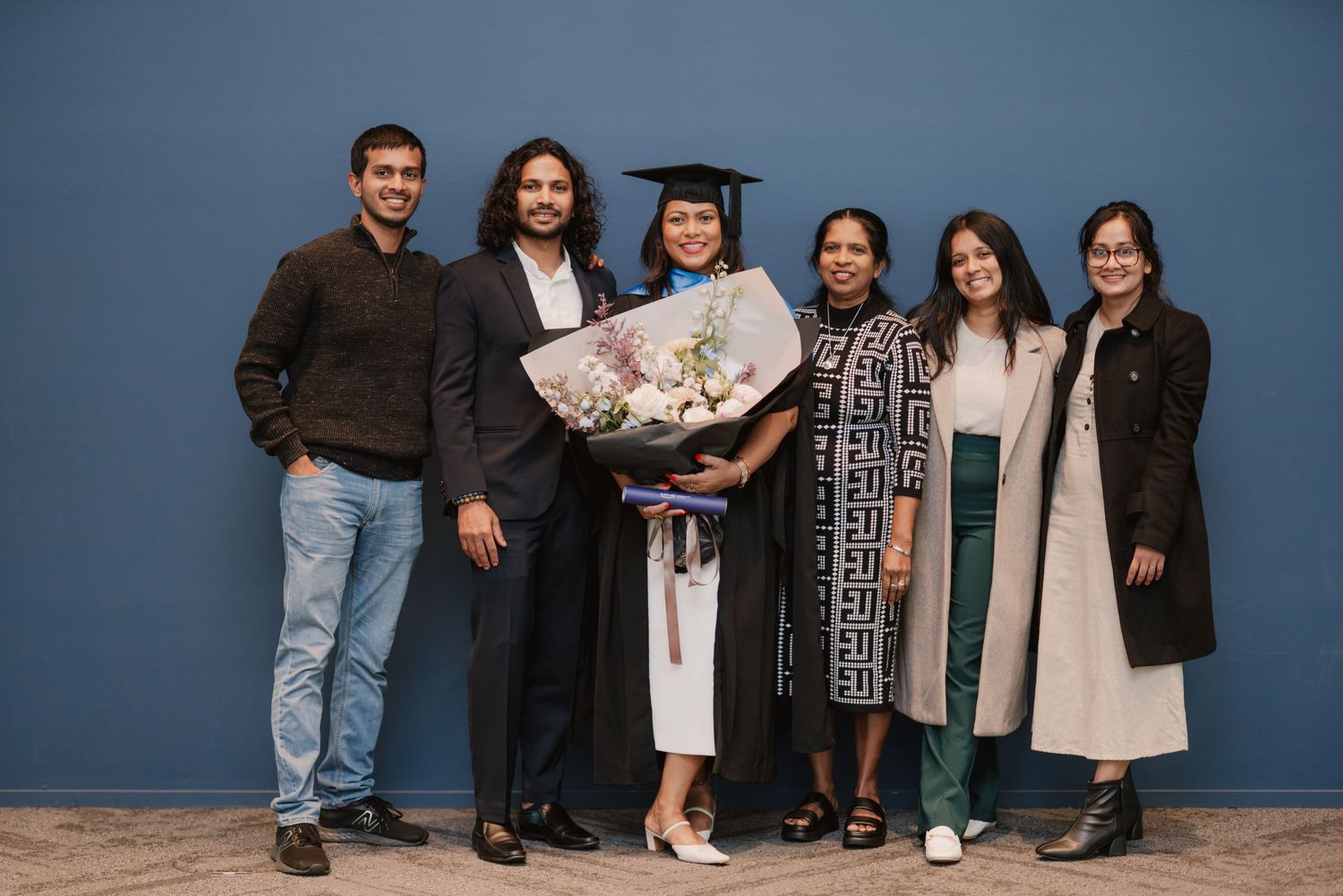 Group of six people posing with a female graduate in cap and gown against a blue wall.