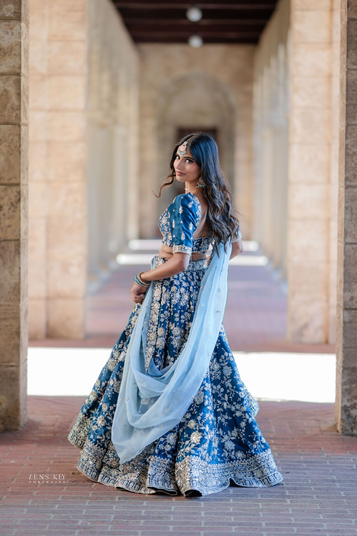 Woman in blue embroidered lehenga with dupatta looking back in columned archway corridor.