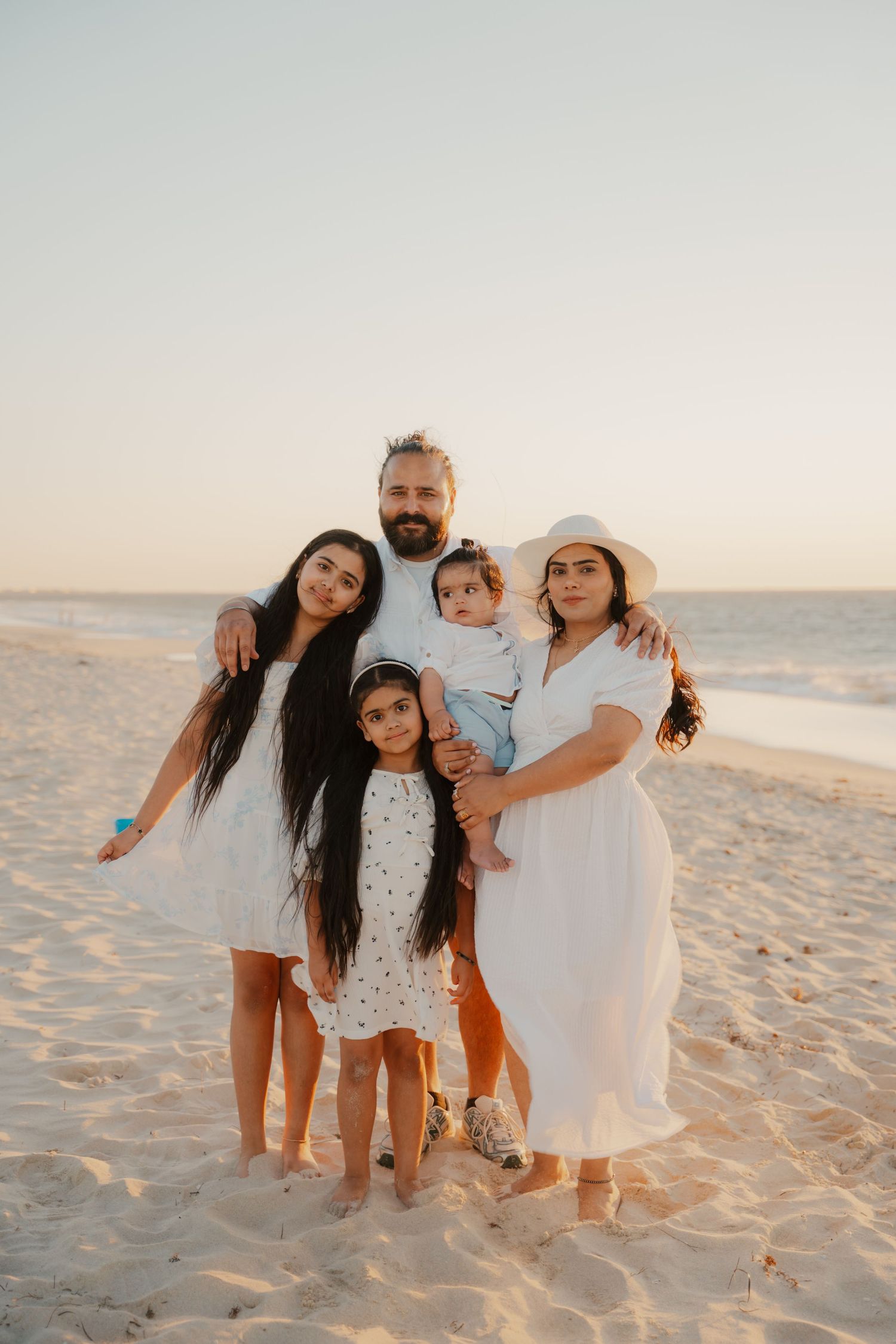 Family of five in white outfits posing together on sandy beach during golden hour sunset.