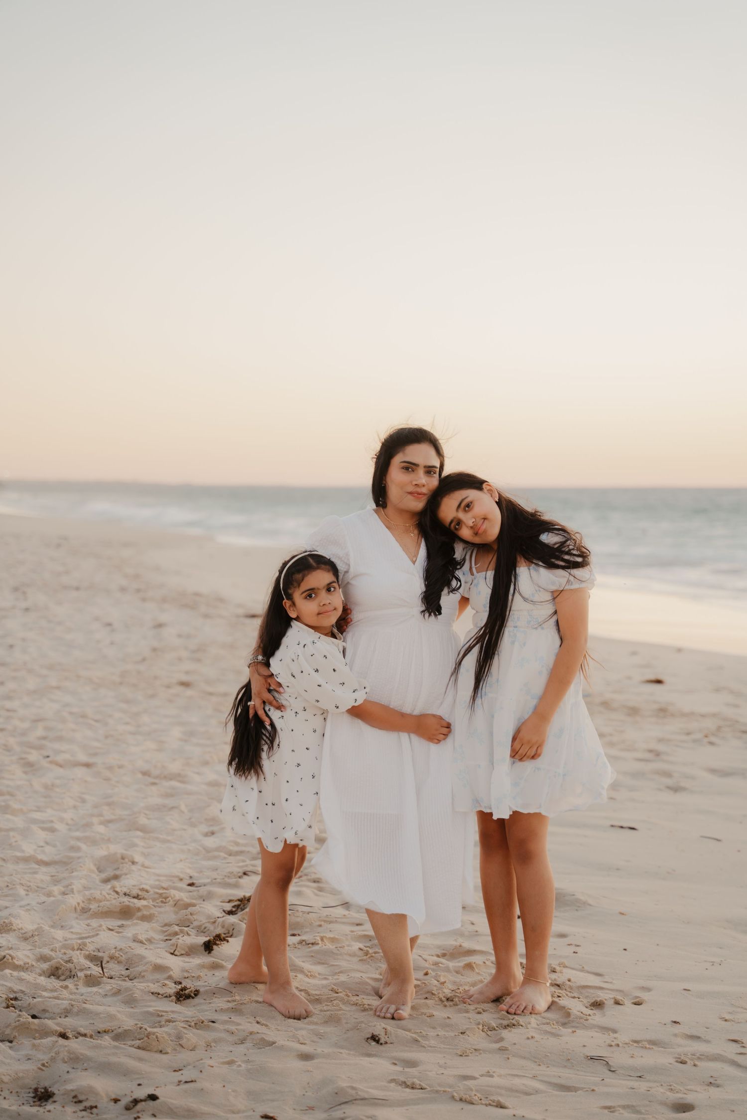 Mother and two daughters in white dresses embracing on sandy beach at dusk.