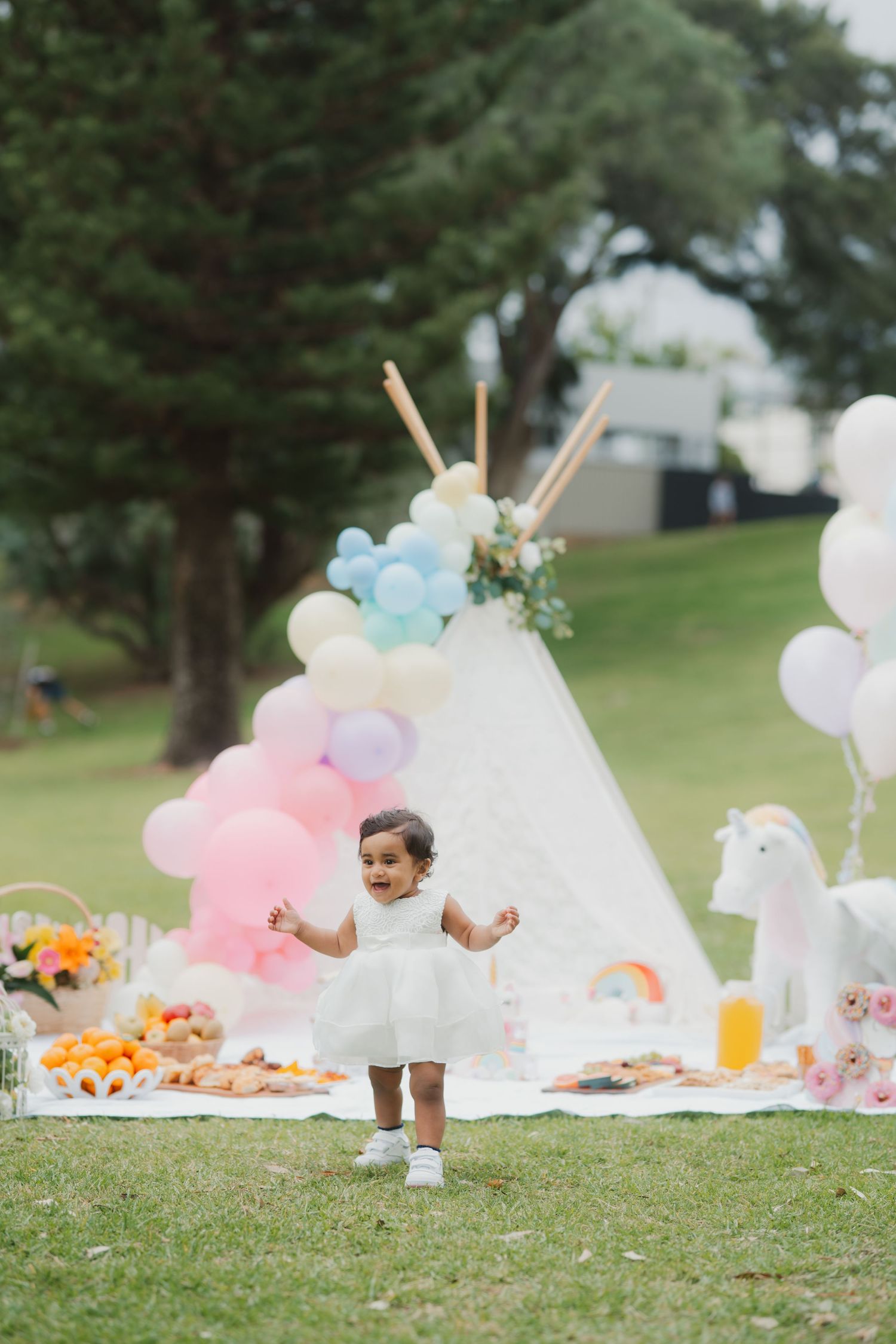 Toddler in white dress laughing at a pastel balloon and unicorn themed outdoor birthday party.