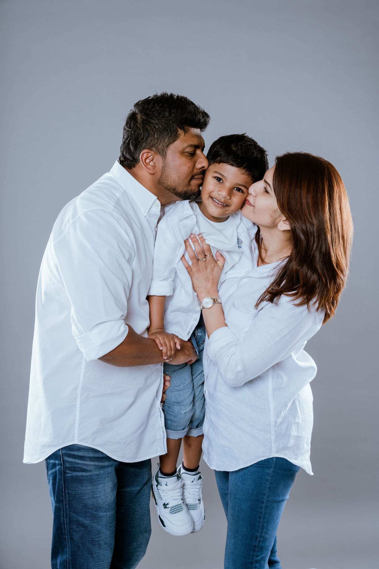 Parents in white shirts kiss their smiling toddler son on a neutral gray background.