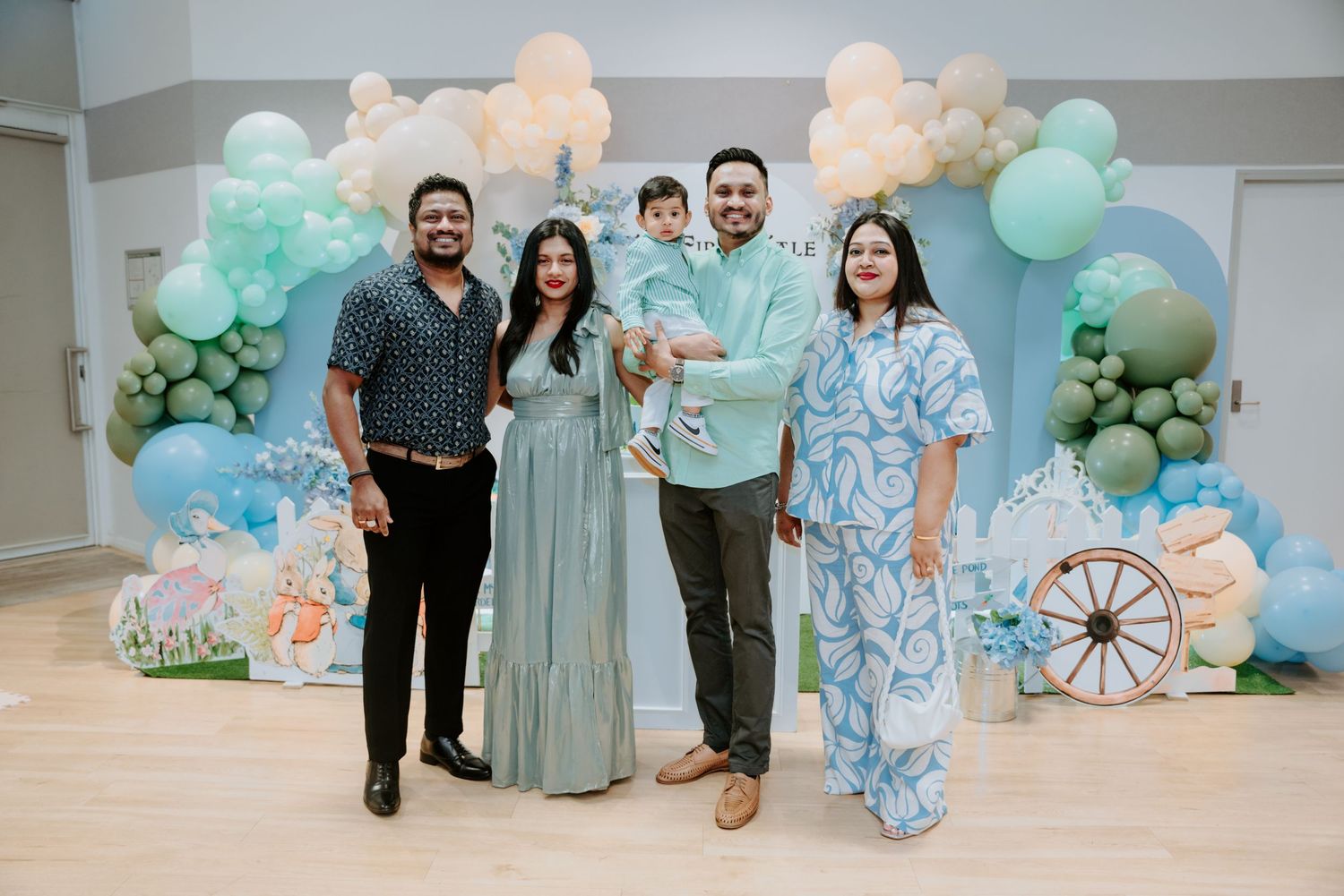 A family of five poses in front of a blue balloon arch at a first birthday party.