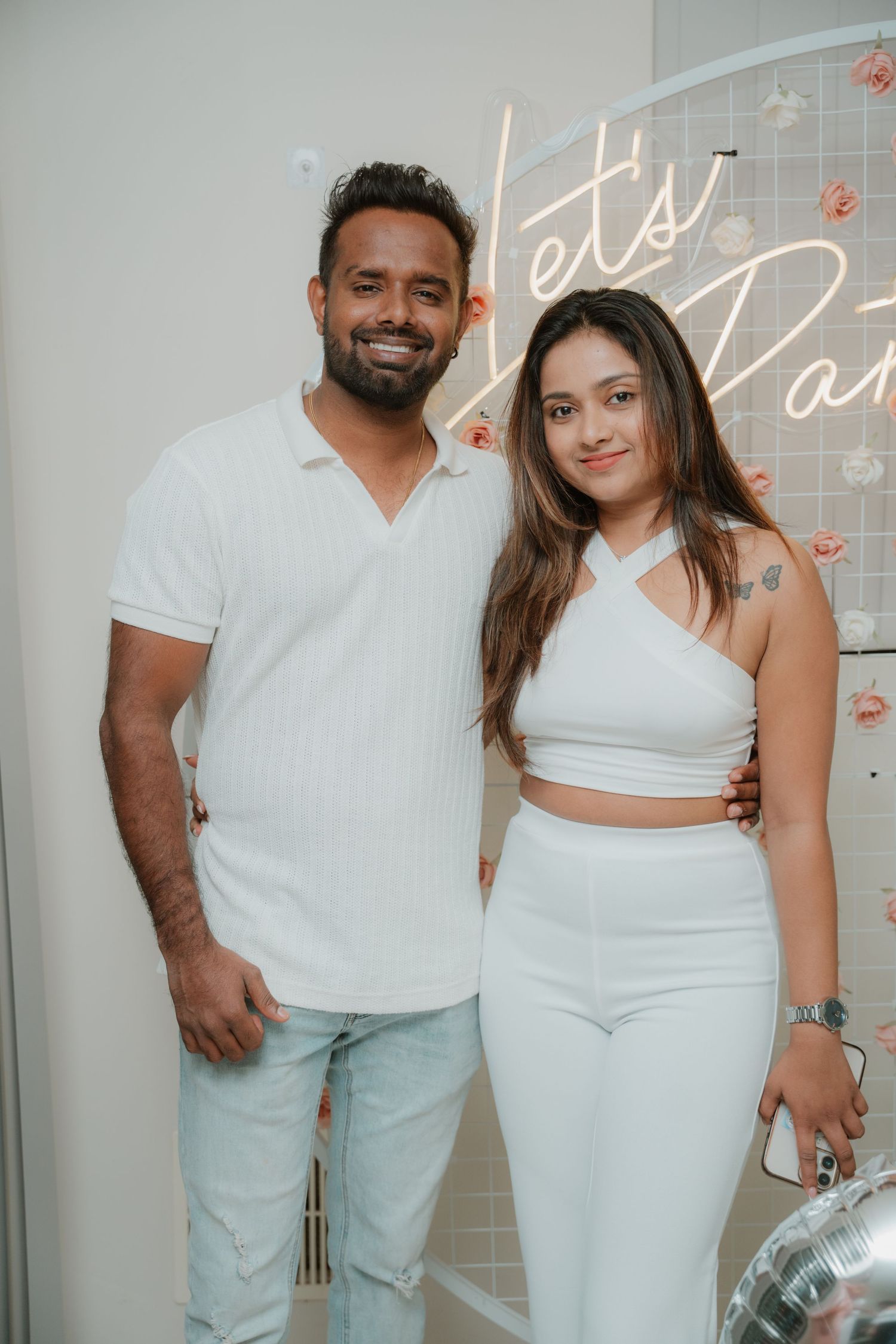 Couple dressed in matching white outfits posing together at a Let's Party neon sign party event.