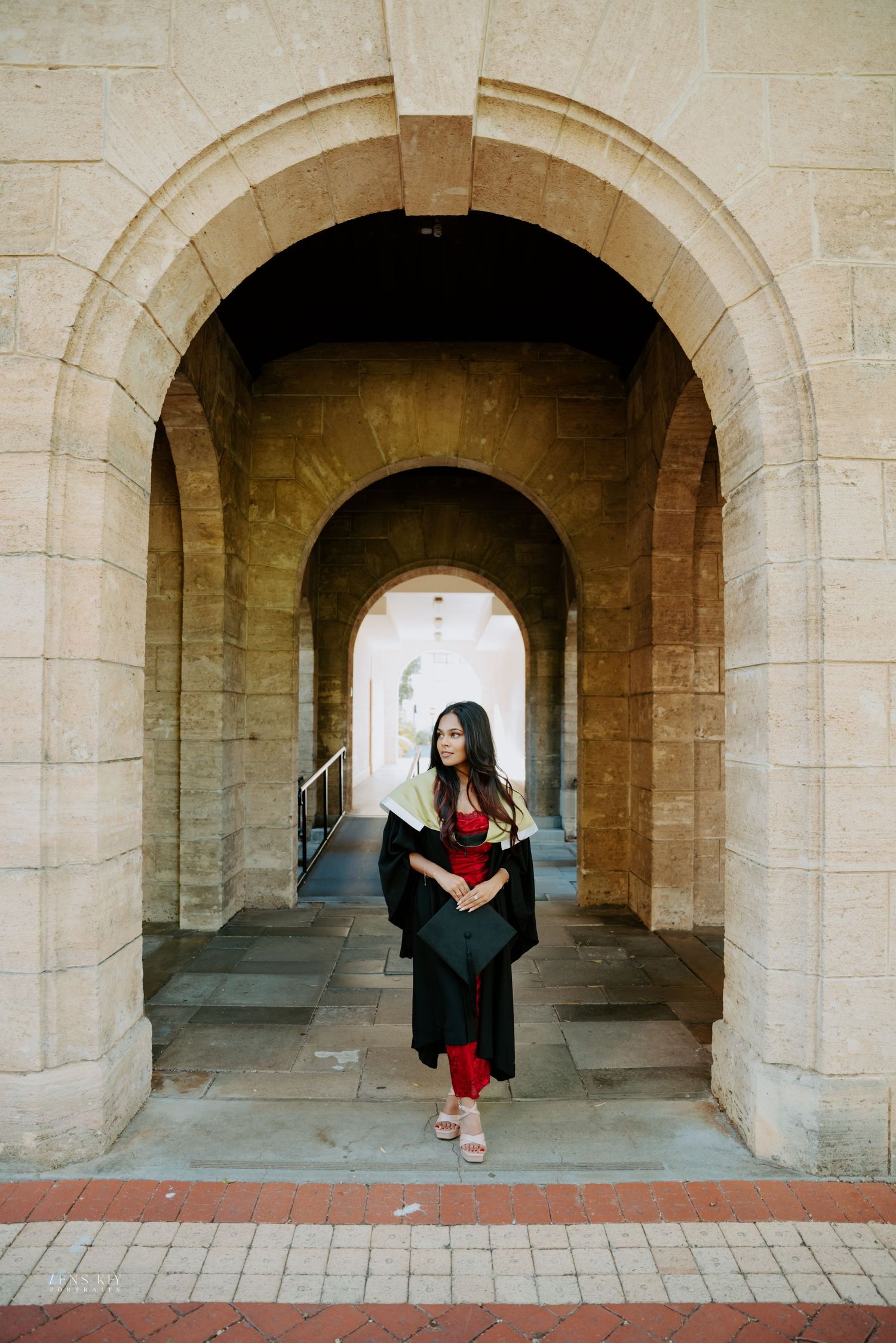 Graduate in black gown and red dress poses under stone archway at university building.
