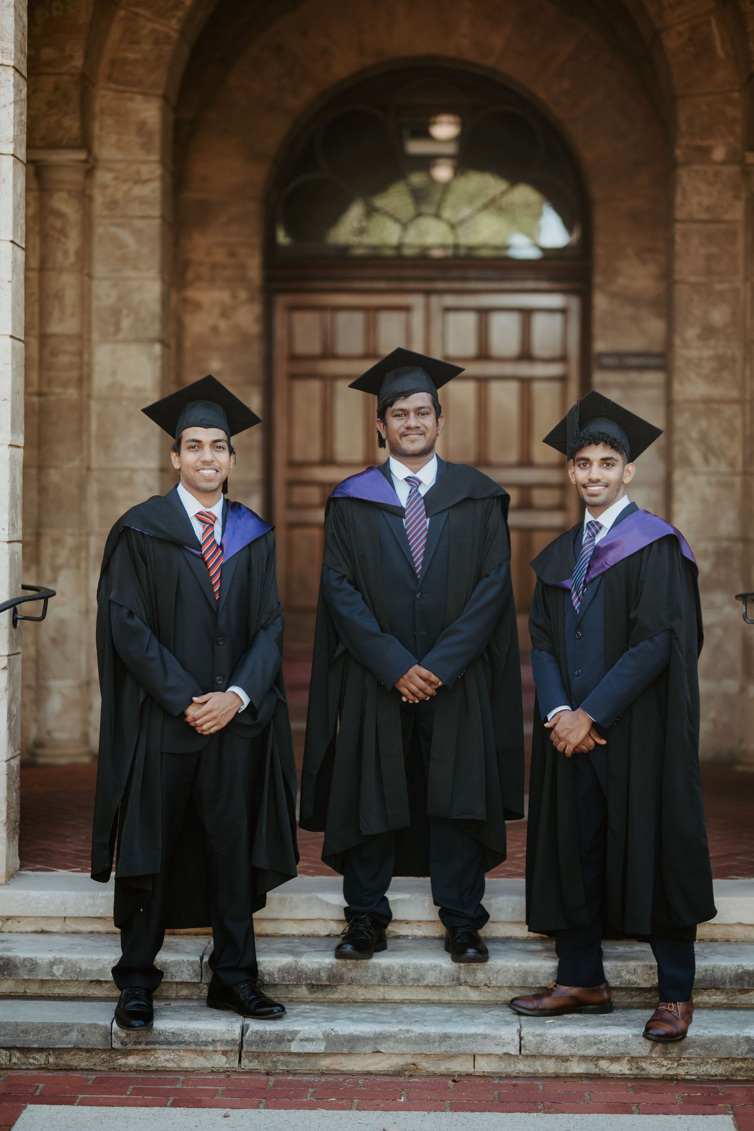 Three male graduates in black caps and gowns with purple hoods posing on university steps.