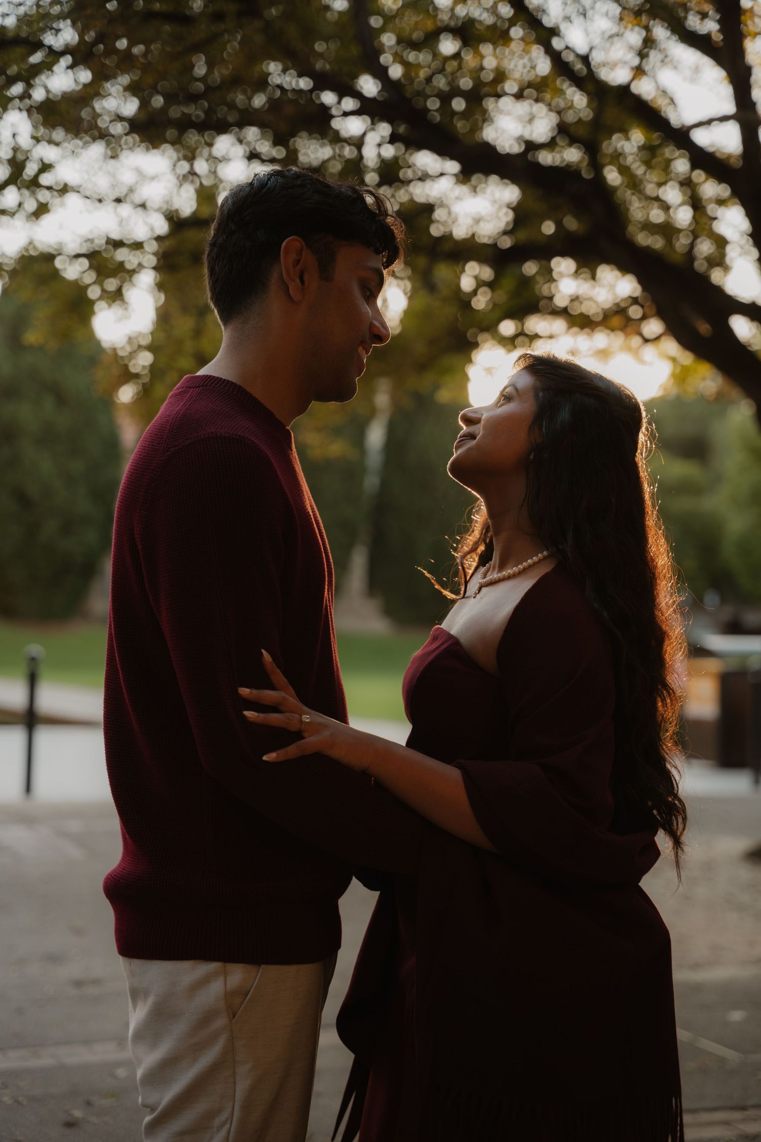 A couple in matching burgundy outfits share a romantic moment outdoors at sunset beneath autumn trees.