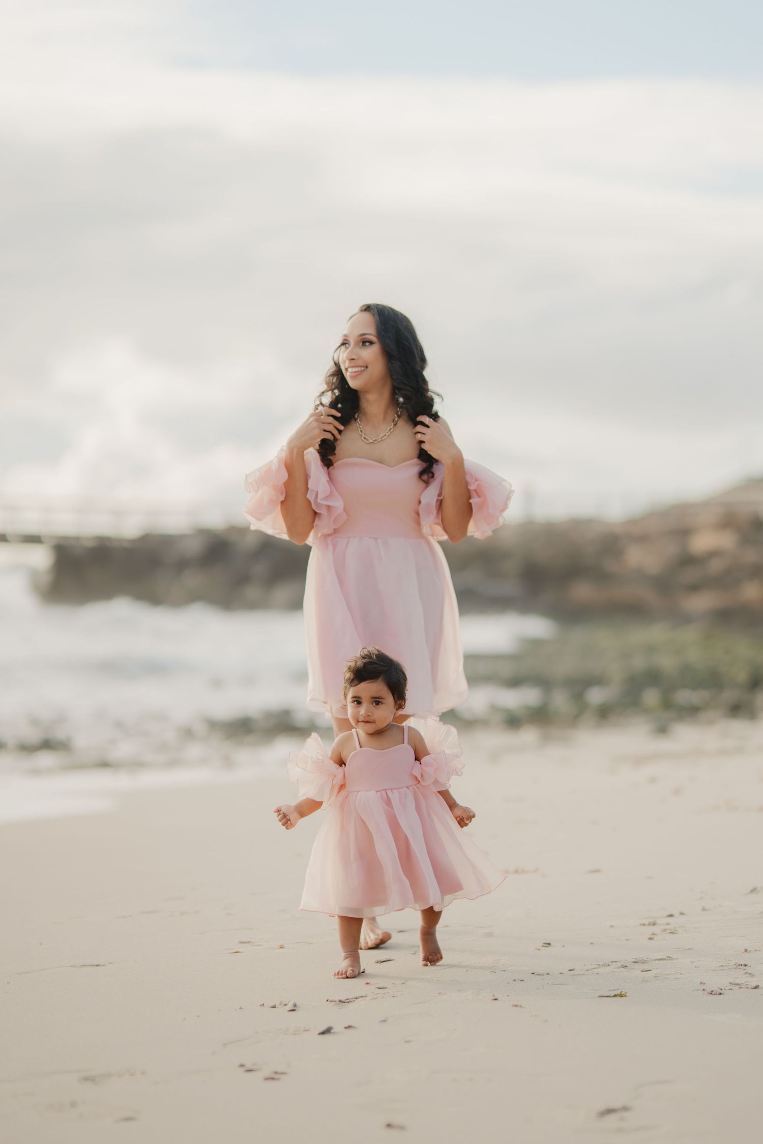 Mother and toddler daughter in matching pink dresses walking on beach together.