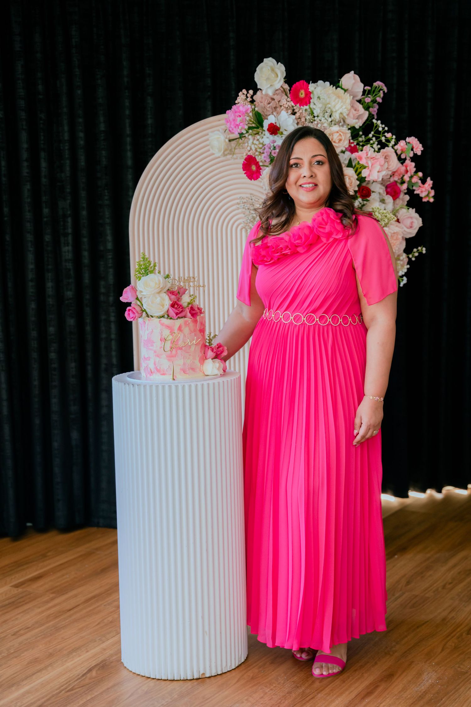 Woman in hot pink pleated gown poses beside floral birthday cake at elegant celebration event.
