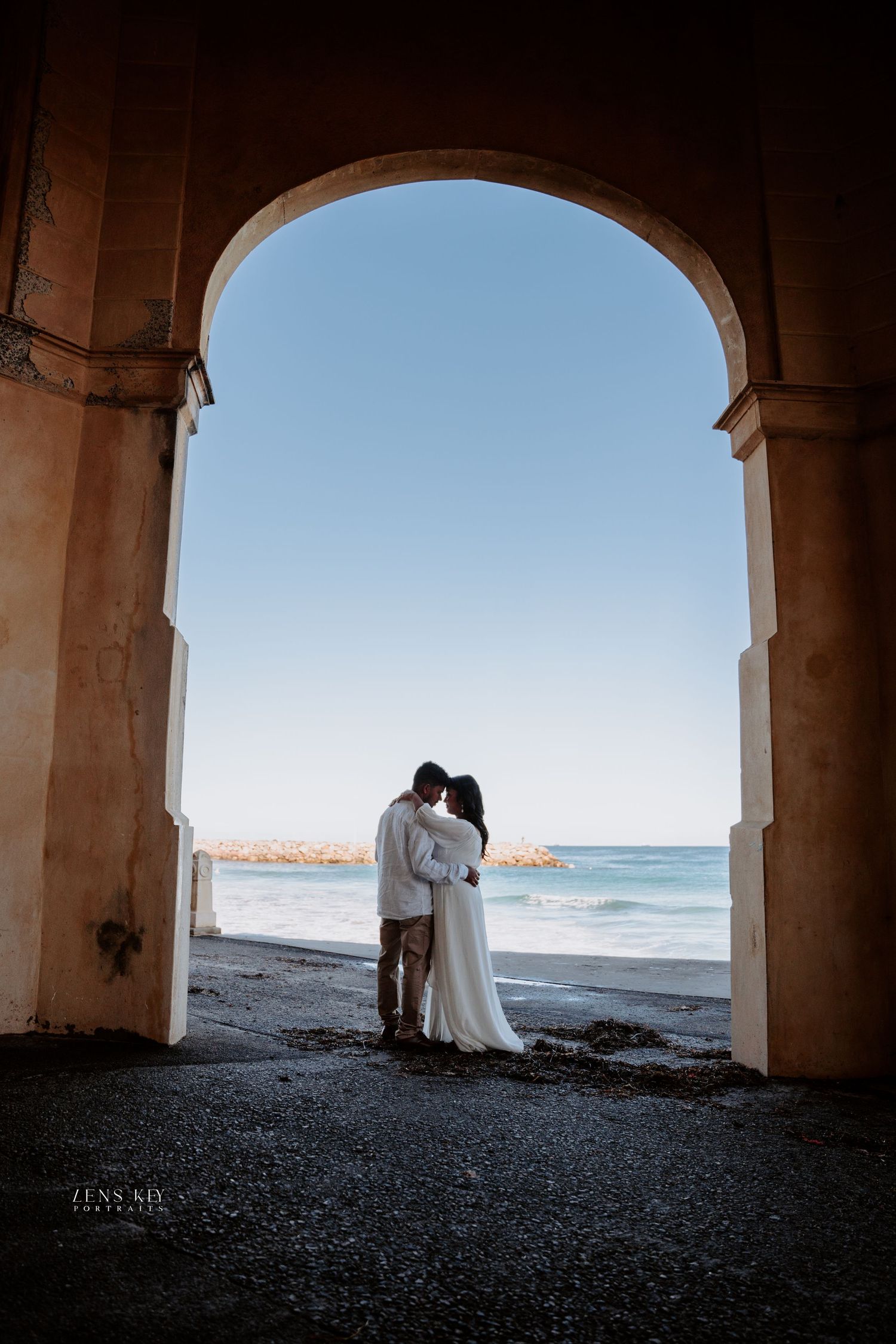 Couple silhouetted in romantic embrace framed by an arched doorway with ocean and blue sky backdrop.