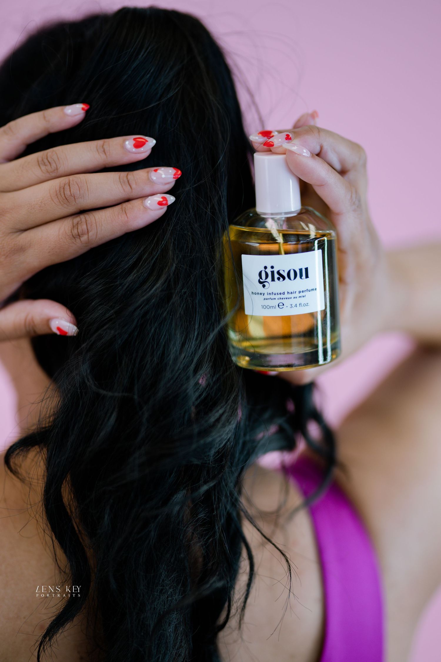 Woman with dark wavy hair applying Gisou honey infused hair perfume spray against pink background.