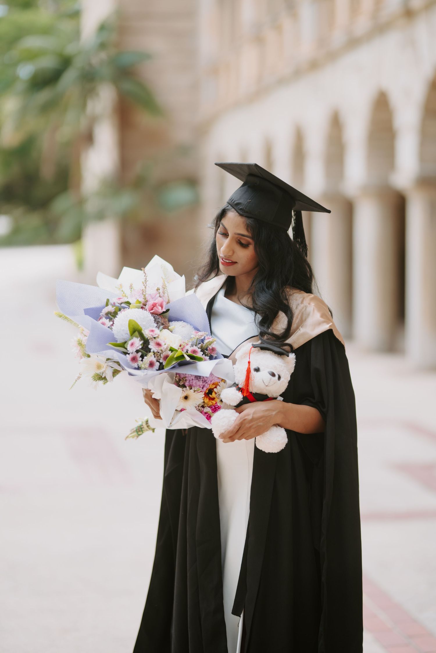 Female graduate in black cap and gown looking down at a bouquet of flowers and teddy bear.