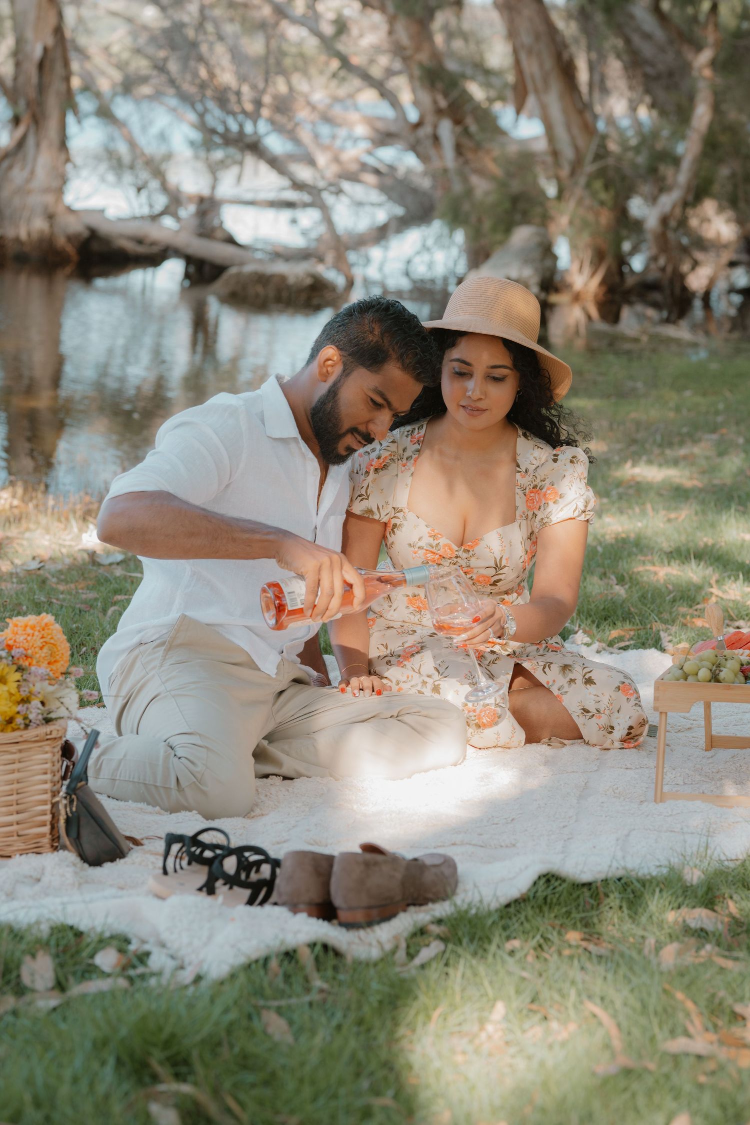 Couple sits on picnic blanket by lake as man pours champagne in golden sunset light outdoors.