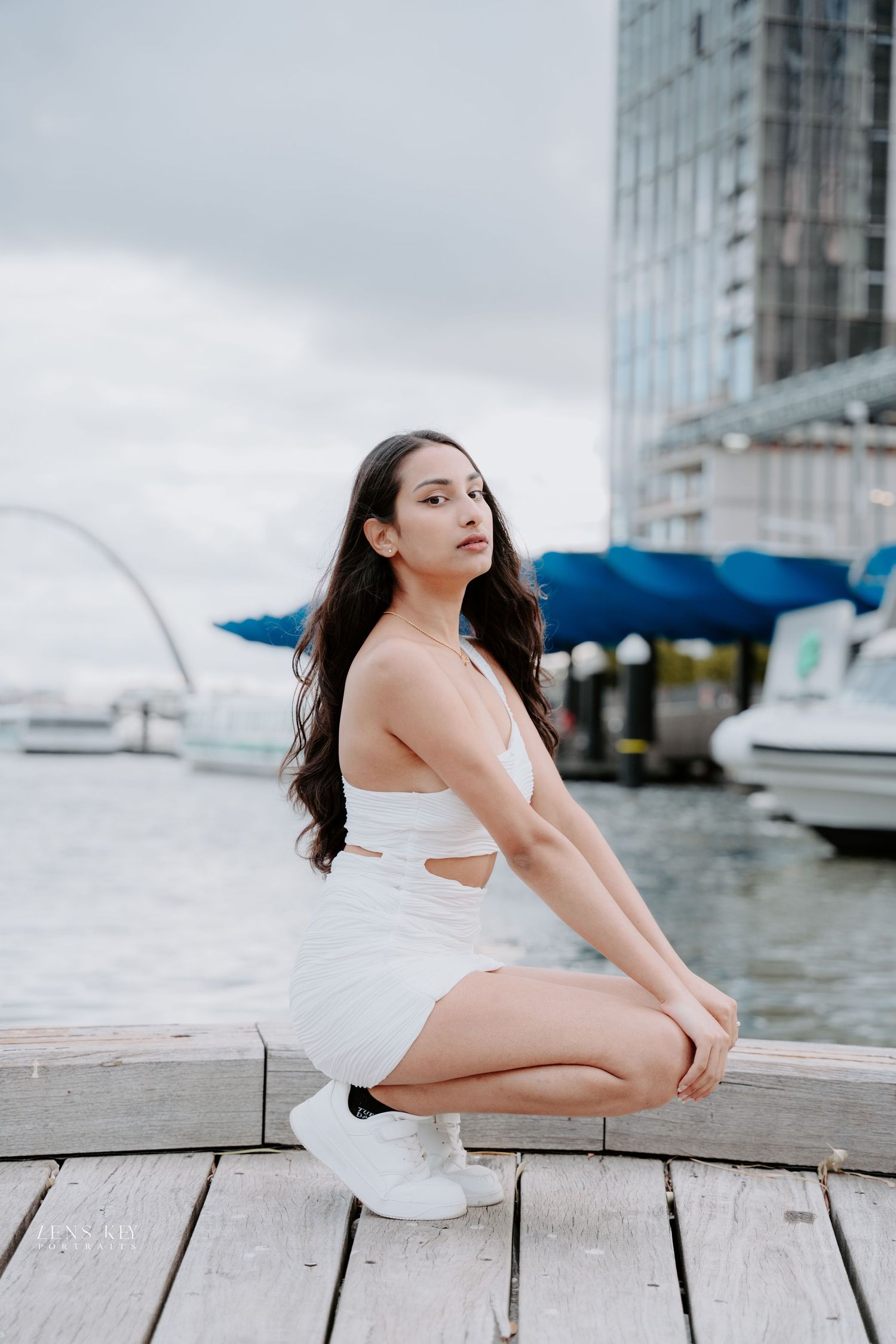 Young woman in white cutout dress crouches on wooden dock with marina and city skyline behind her.