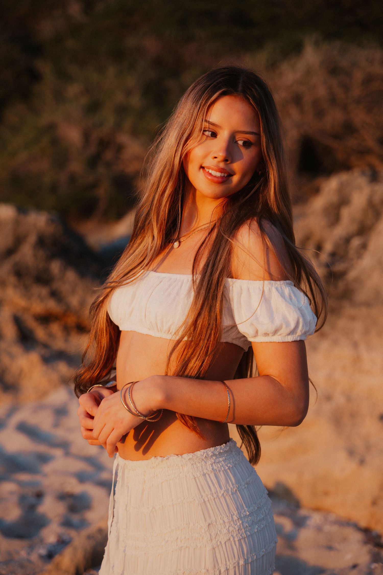 Smiling young woman in white off-shoulder crop top and skirt posing on sandy beach at sunset.