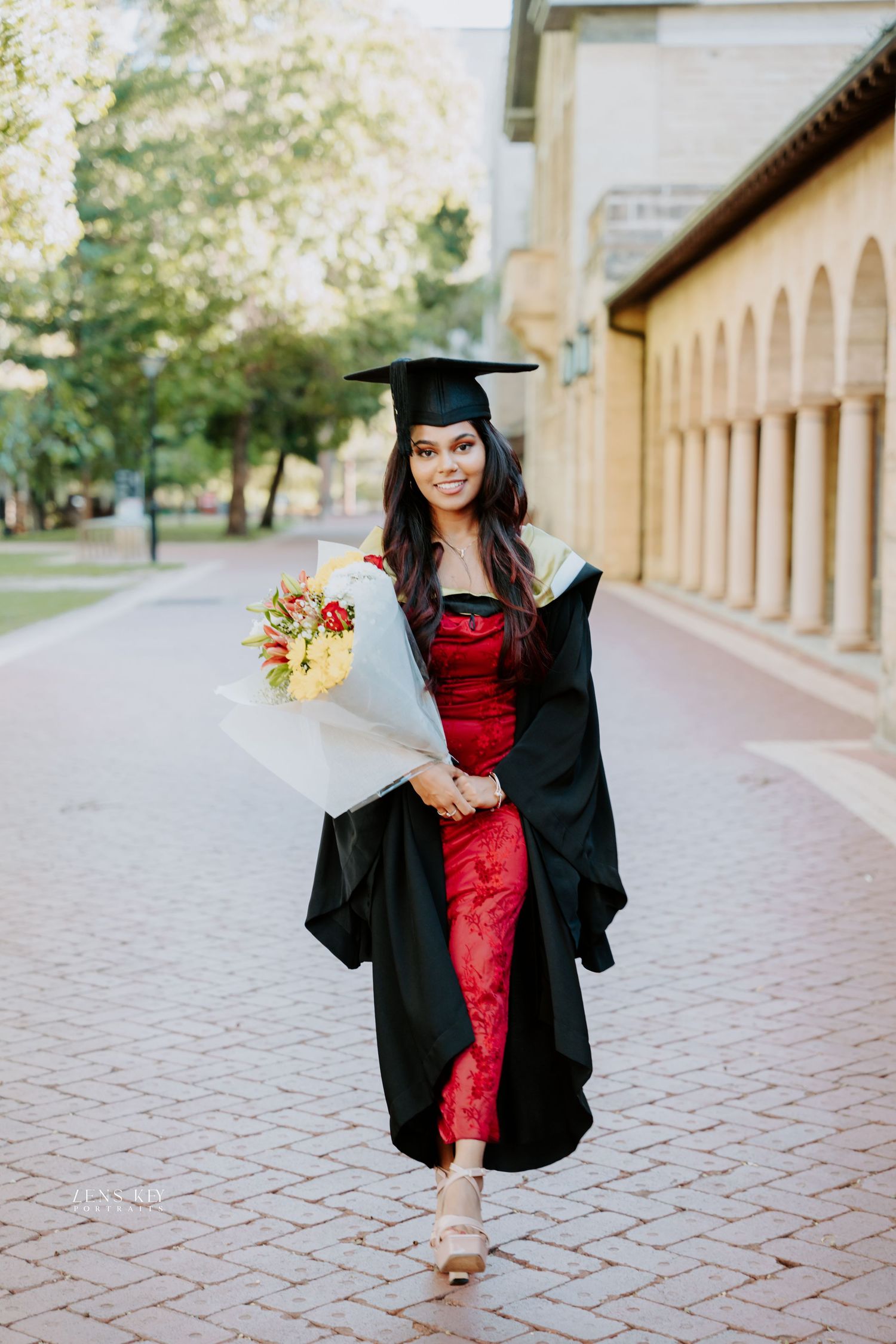 Smiling female graduate in red dress and black gown walks holding a bouquet of flowers on campus.
