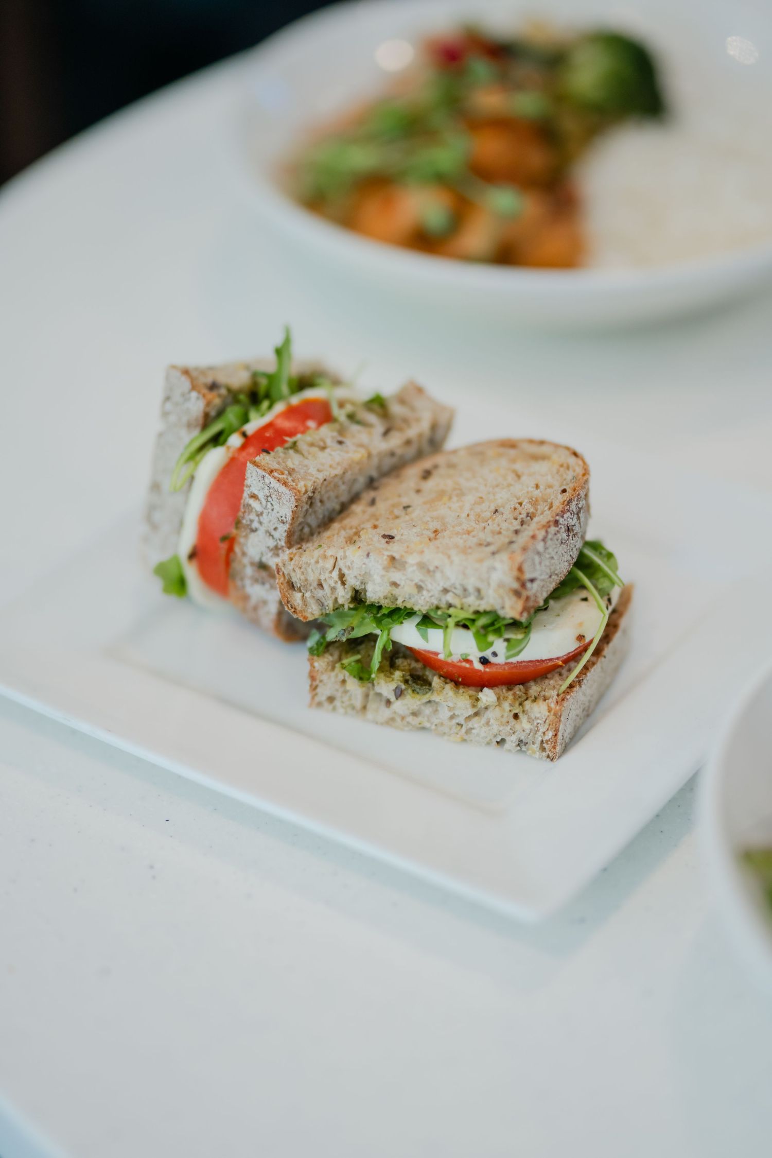 A fresh vegetable sandwich with tomato, mozzarella, and greens on multigrain bread on white plate.