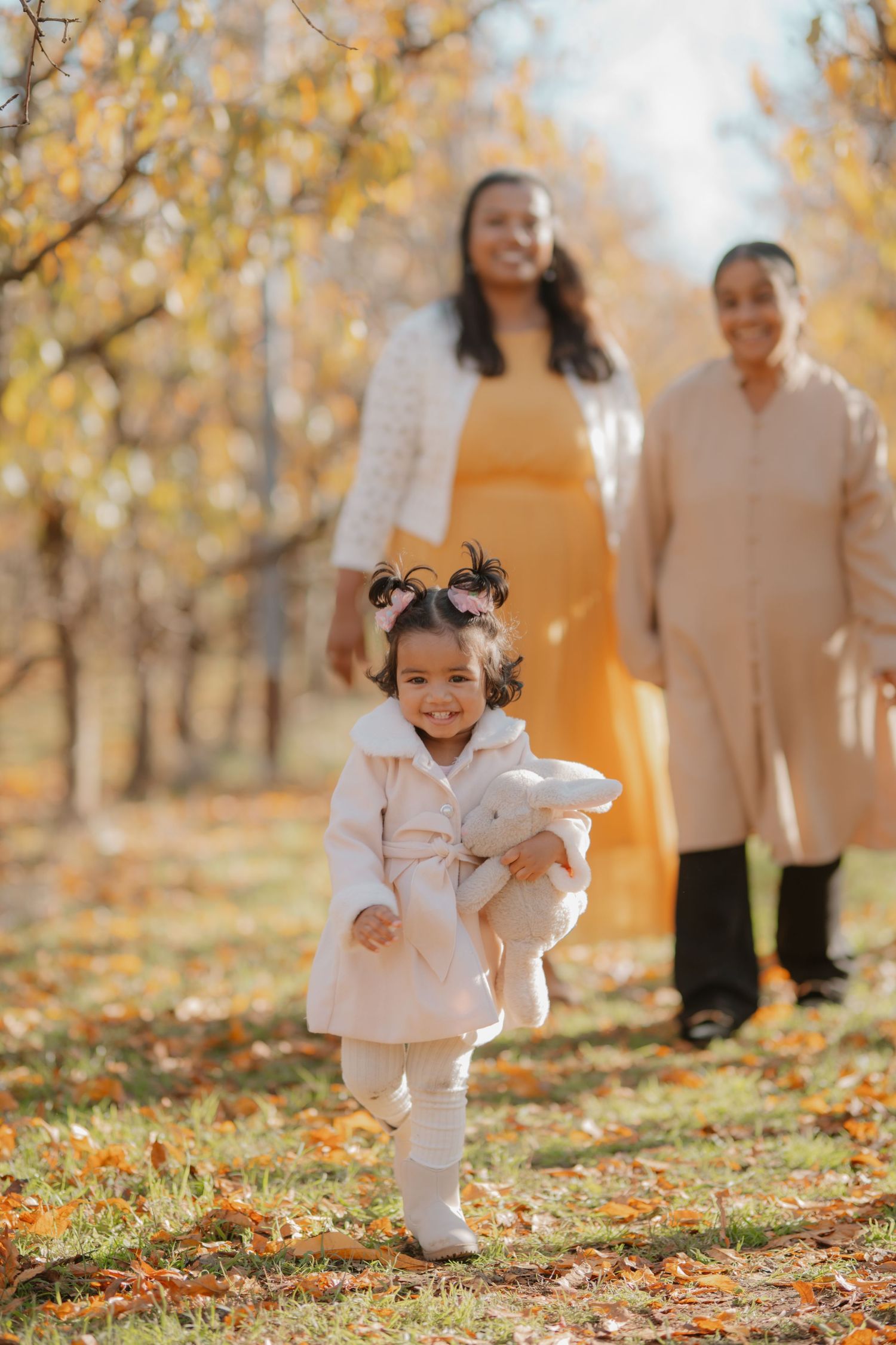Toddler girl in pink coat runs happily toward camera as mother and grandmother walk behind in autumn park.