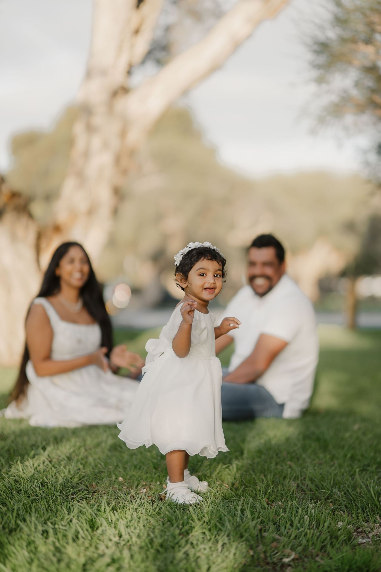 Toddler girl in white dress playing on green grass while parents smile in the background.