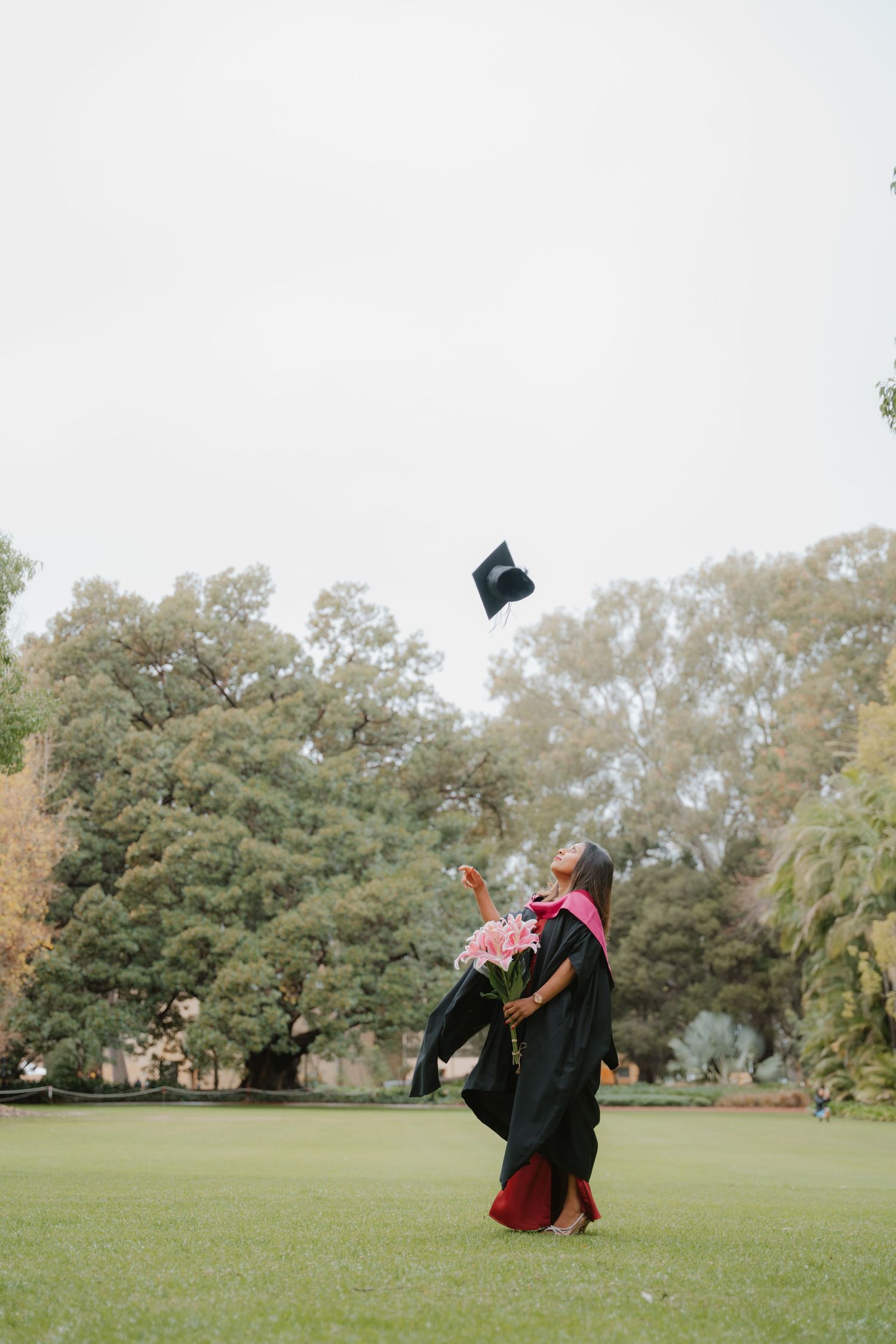 Graduate in black gown tosses mortarboard cap in the air on green campus lawn.
