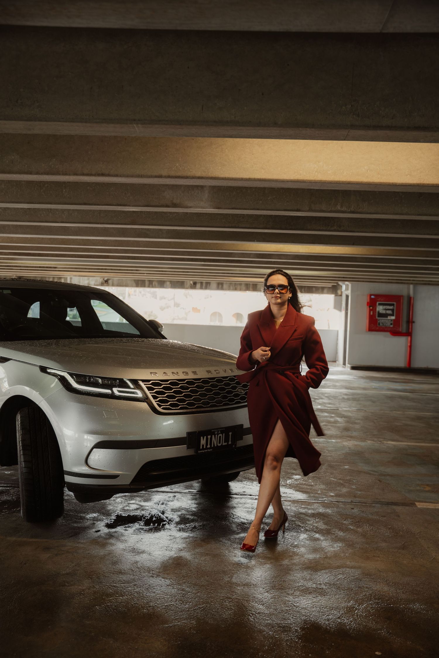Stylish woman in burgundy coat and heels walking beside silver Range Rover Velar in parking garage.