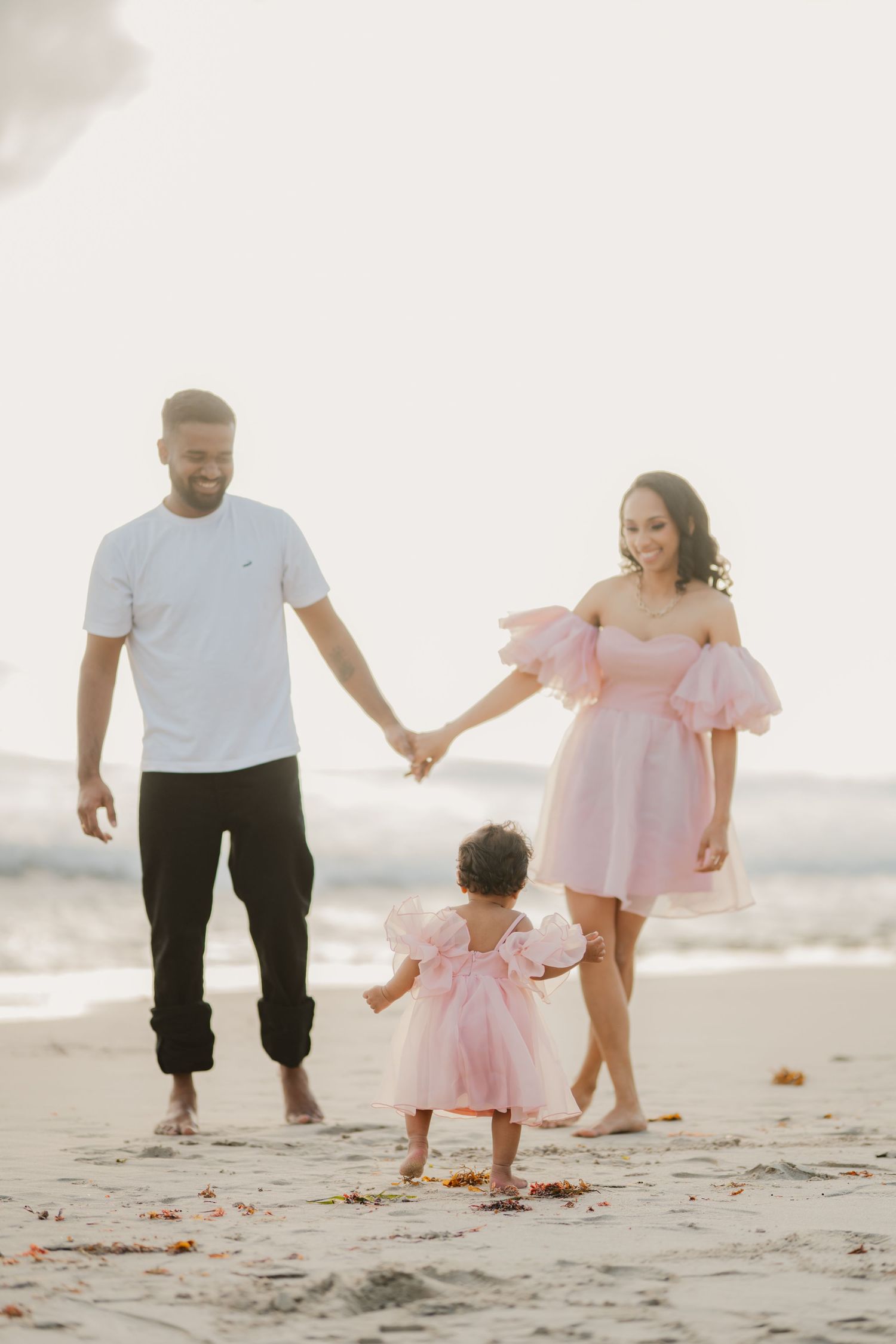 Family of three in pink outfits holding hands walking on sandy beach.
