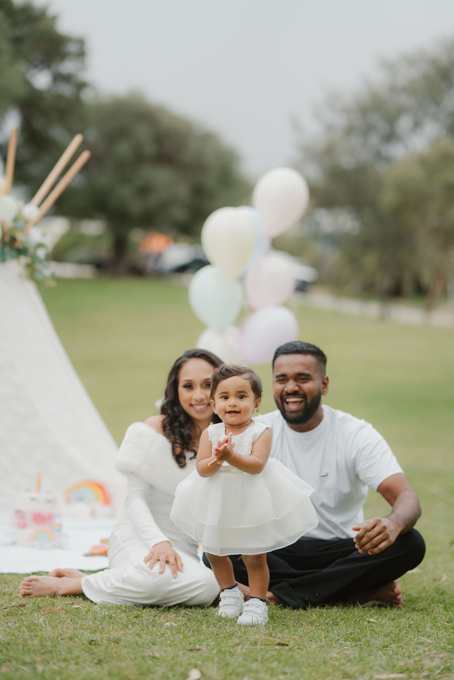 Happy family of three sitting on grass at a pastel balloon teepee first birthday celebration.