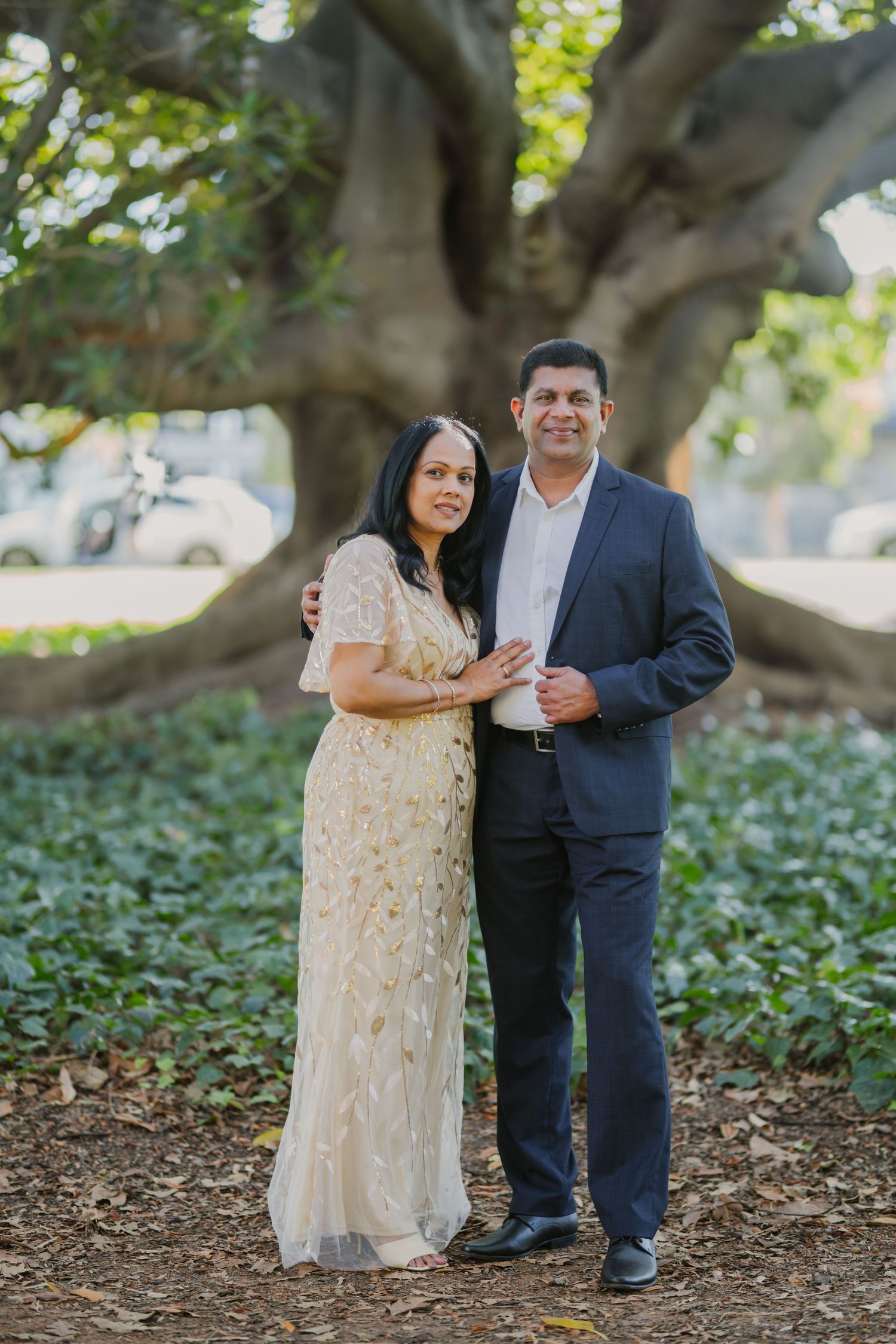 Elegant couple poses together under large tree, woman in gold embroidered gown, man in dark suit.
