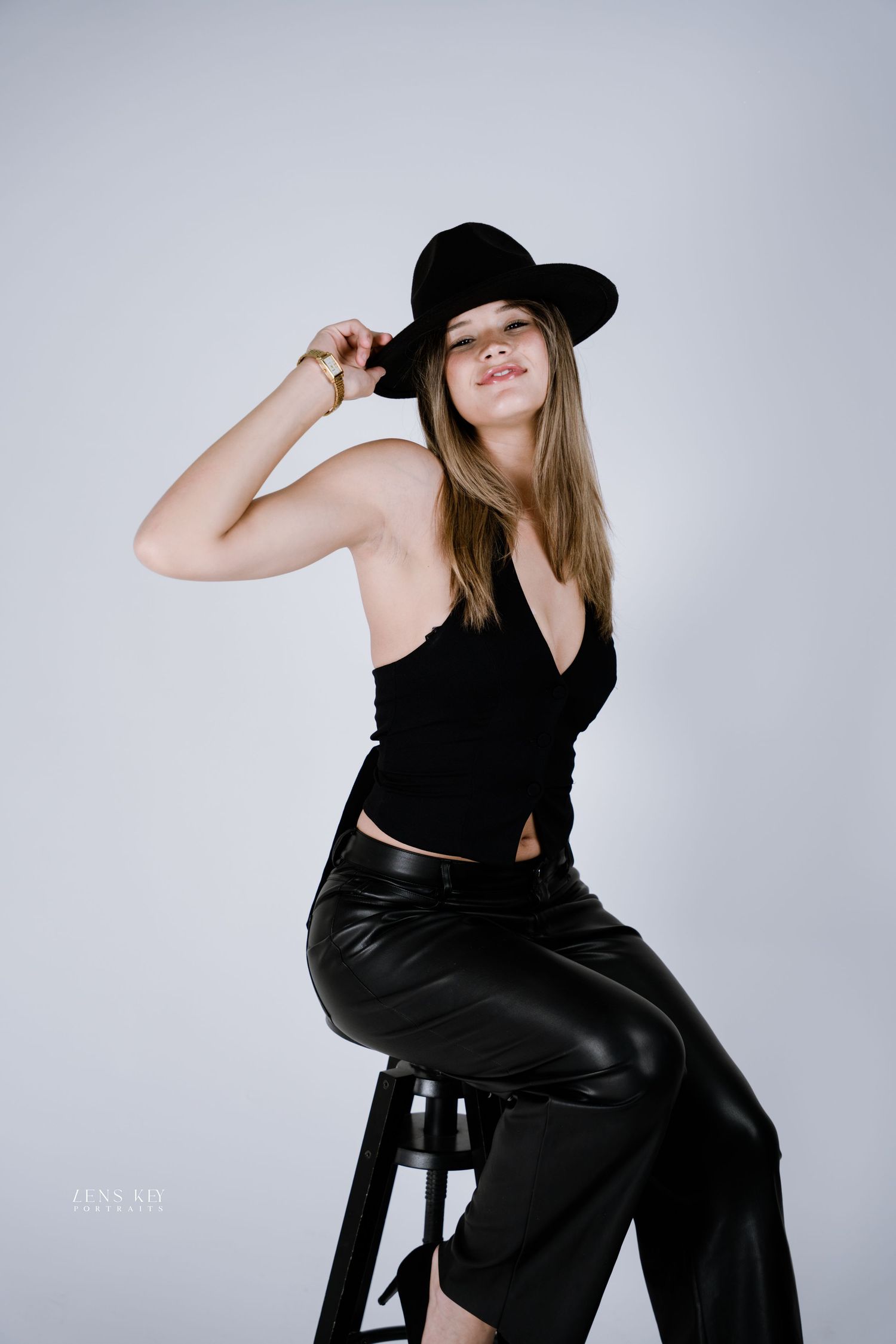 Stylish woman wearing black outfit and wide-brim hat poses seated on stool in white studio.