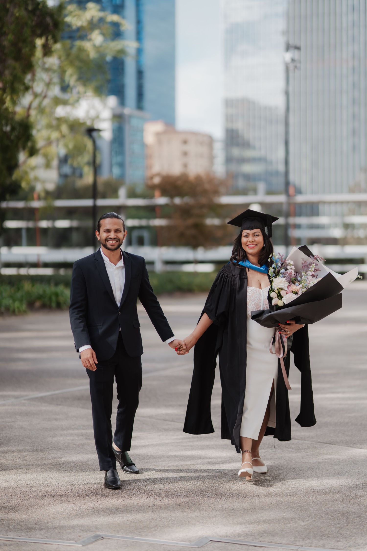 A smiling graduate in cap and gown holds flowers while walking hand in hand with a man outdoors.