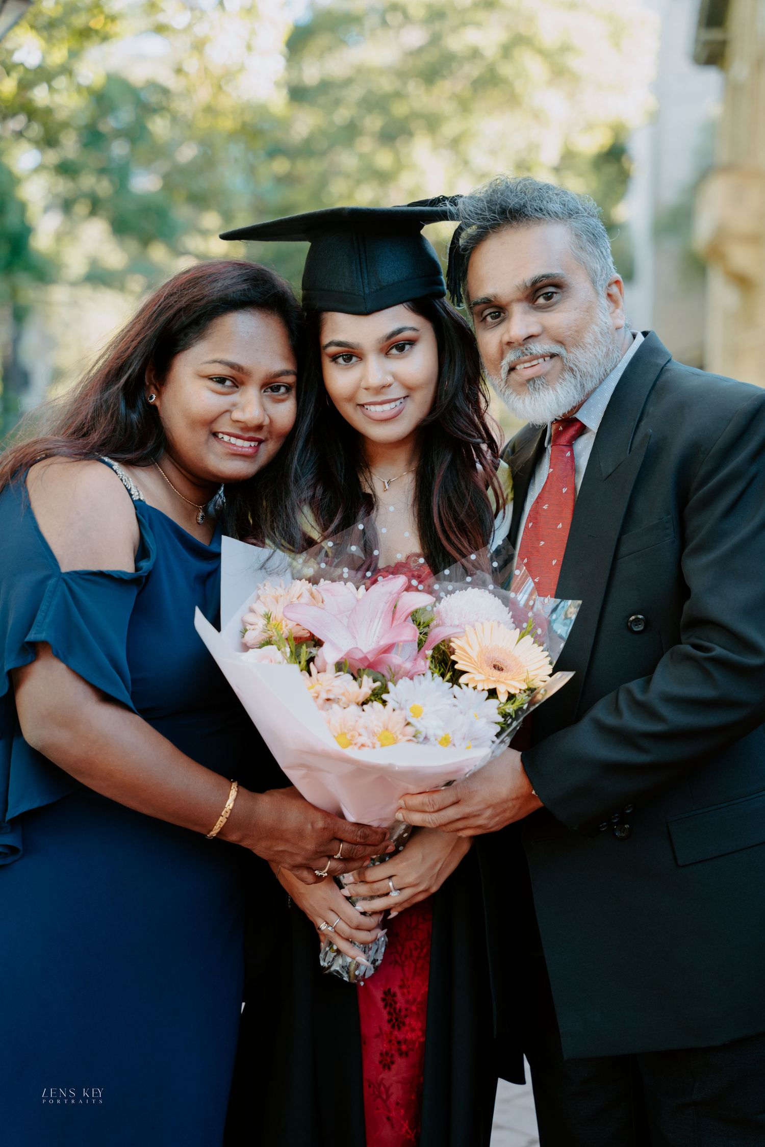 Proud graduate in cap and gown holding bouquet of flowers with her parents at graduation.