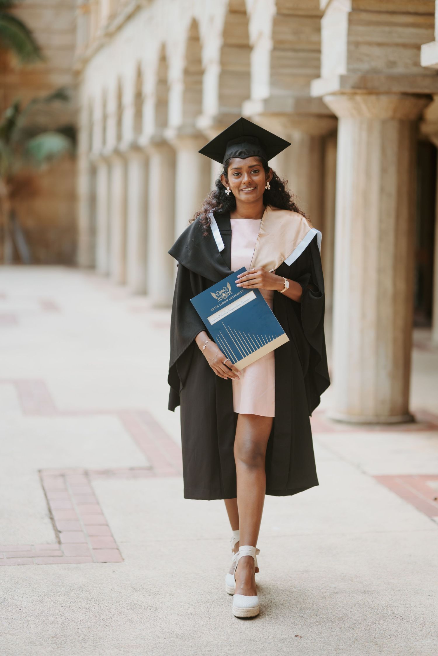 Female graduate in black cap and gown walking and holding a diploma in a columned hallway.