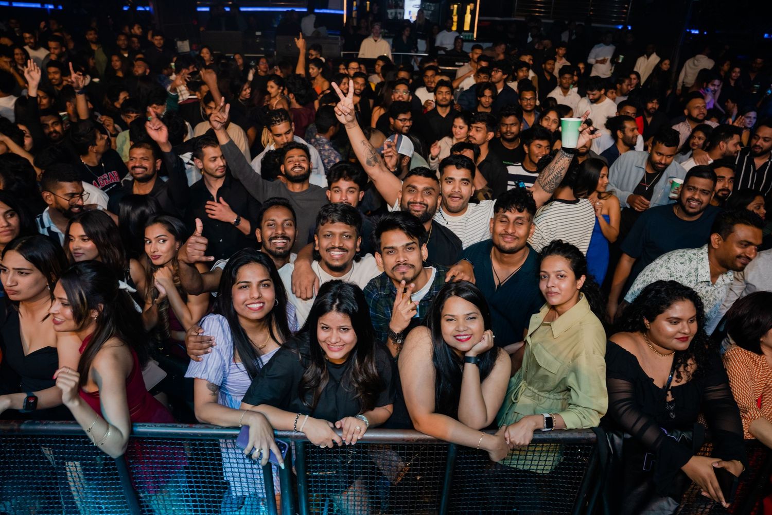 Large diverse crowd of concert attendees smiling and gesturing at an indoor music event.