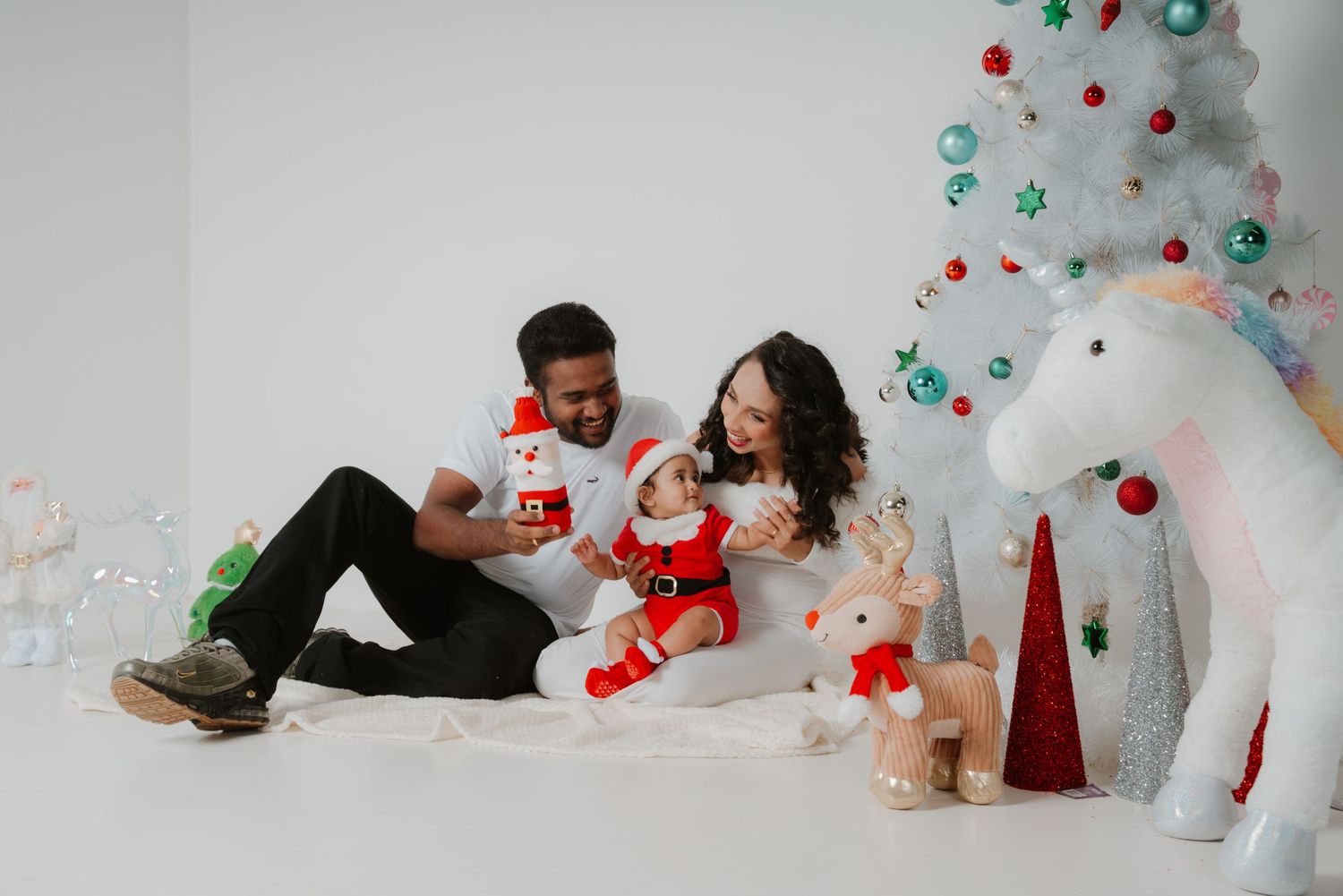 Happy family with baby dressed in Santa outfit celebrating Christmas with festive decorations and white tree.