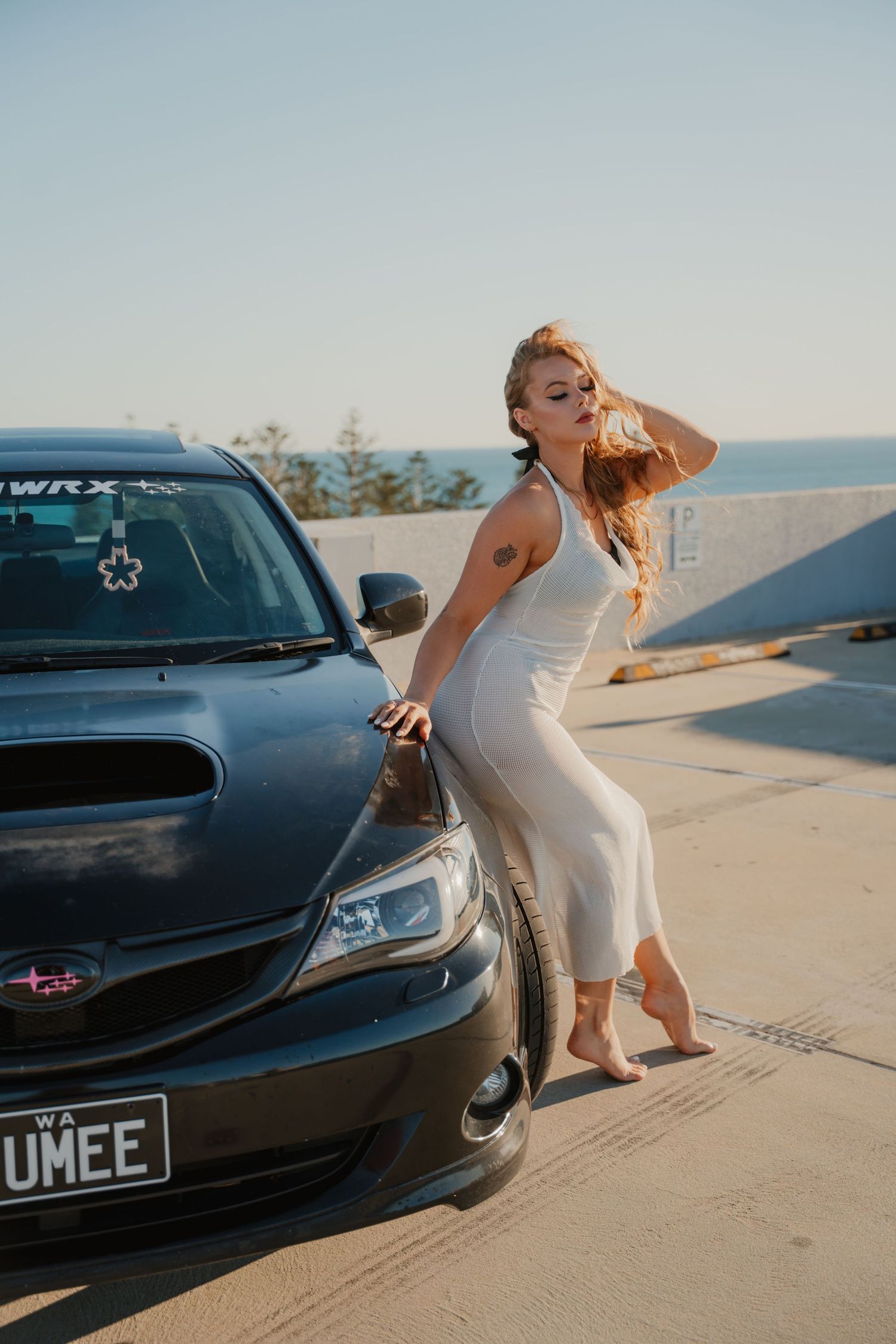 Redhead woman in white dress posing beside black Subaru WRX with ocean view background.