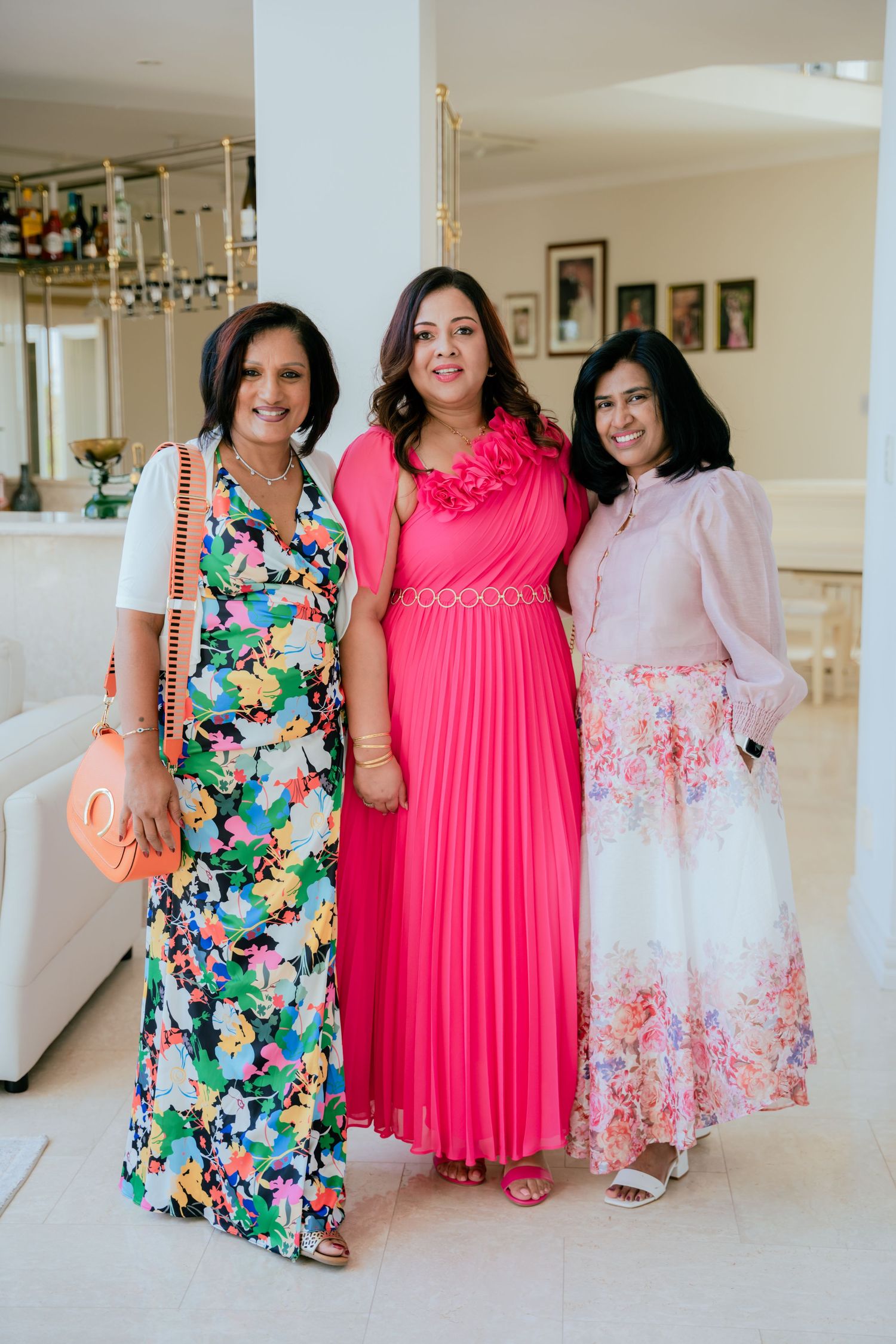 Three women in colorful dresses pose together at an elegant indoor birthday party celebration.