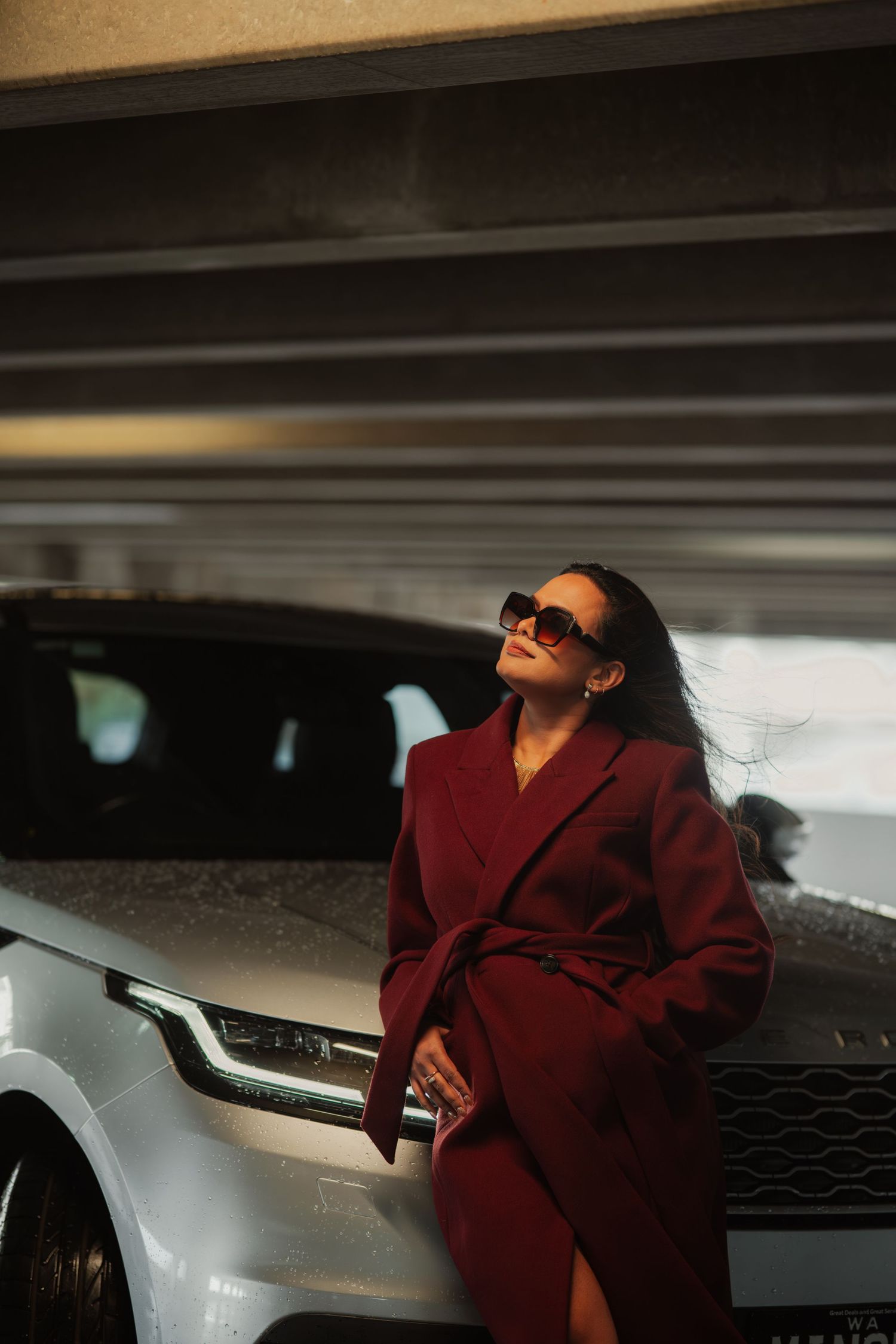 Sophisticated woman in burgundy coat and sunglasses posing beside silver Range Rover in dramatic garage lighting.
