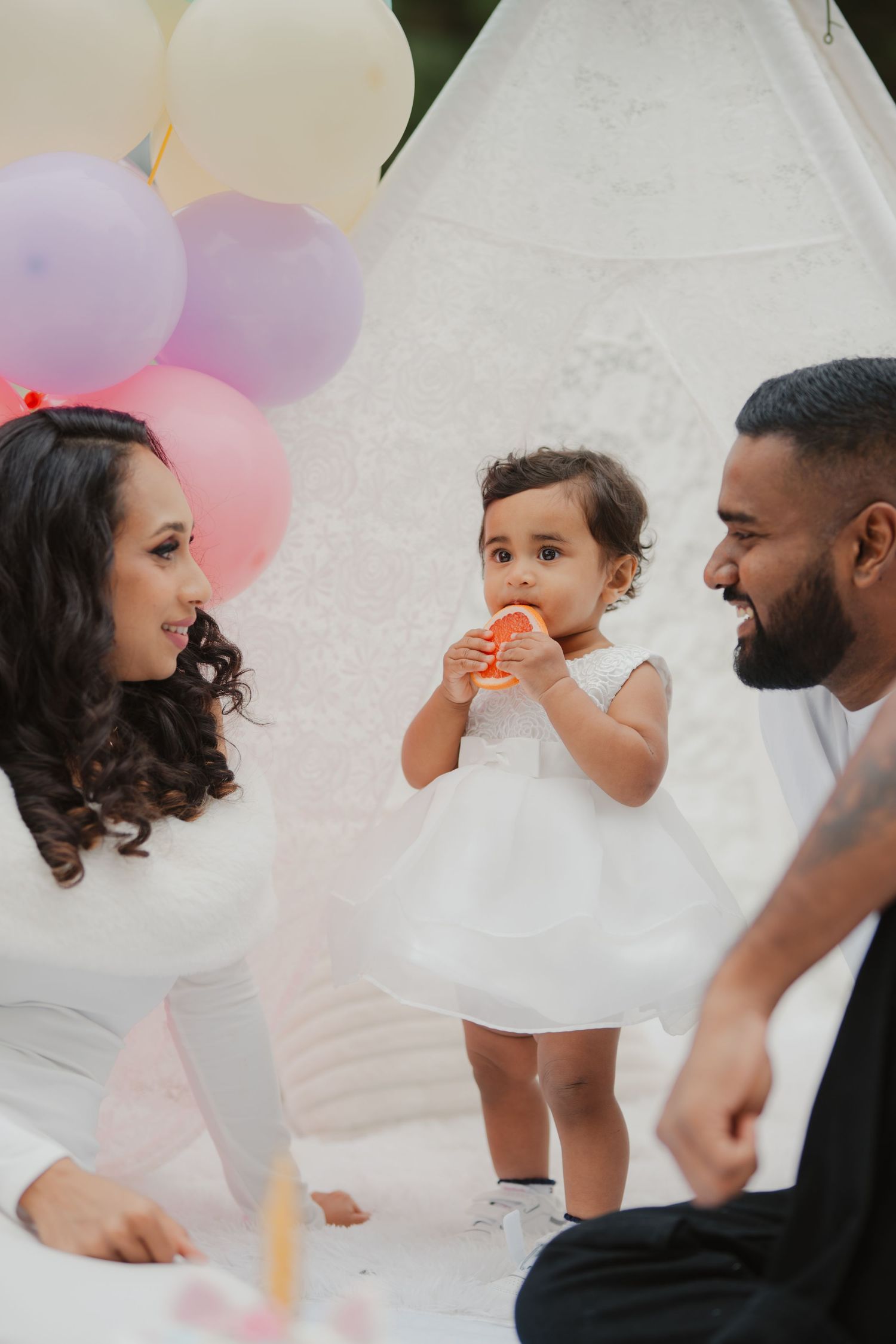 Parents smiling at toddler in white dress eating fruit at colorful balloon-decorated birthday party.