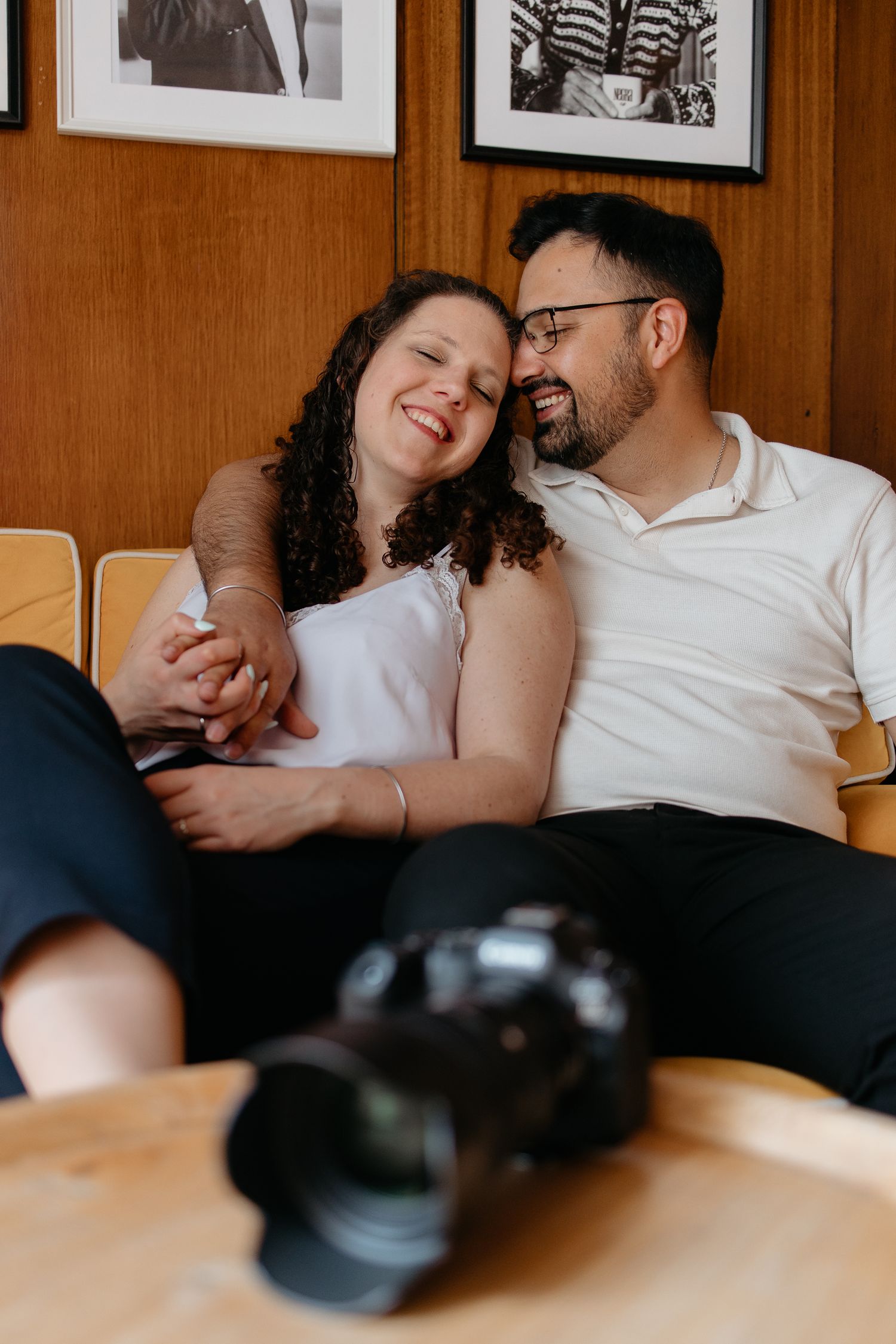 A happy couple sits together on a chair, holding hands and smiling, with a camera in the foreground.