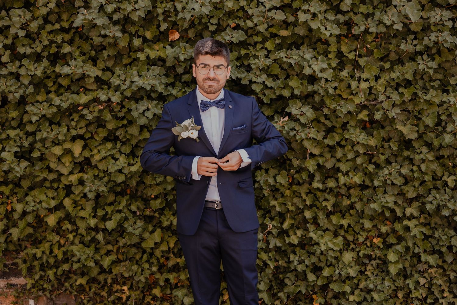 Person in navy suit poses in front of ivy-covered wall outdoors.