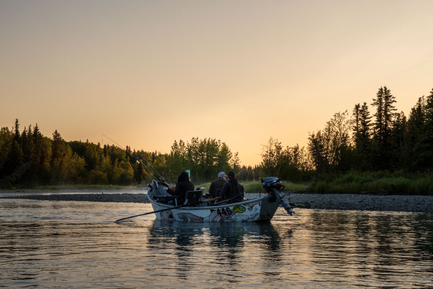 Boats silhouetted against a golden sunset on a tranquil lake with evergreen trees lining the shore.